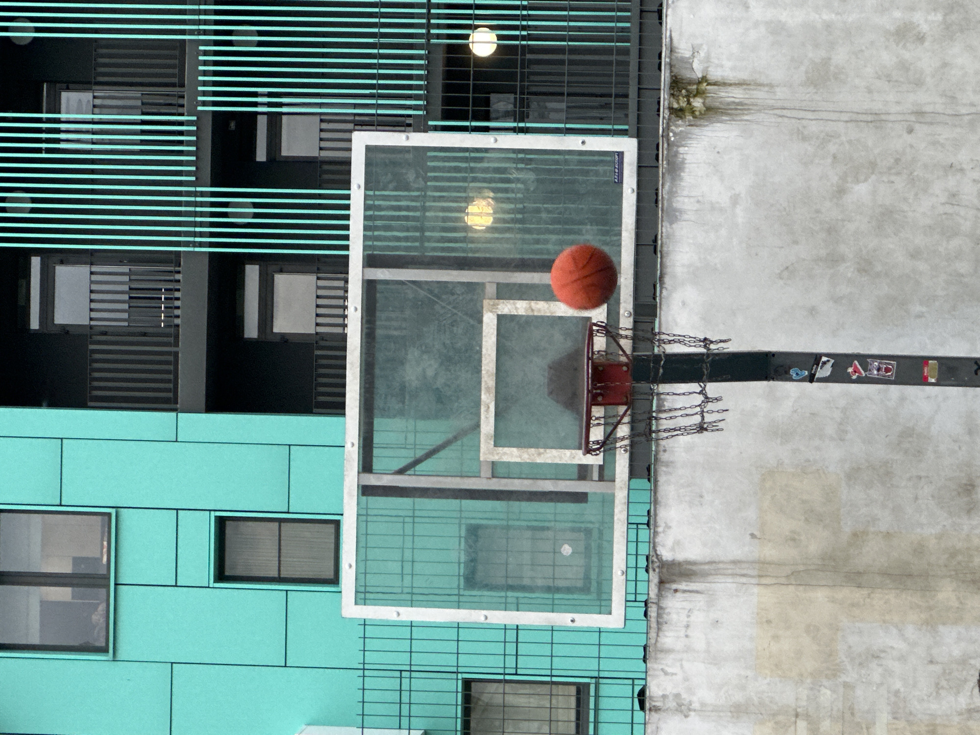 The image shows a basketball hoop with a clear backboard and a red rim, positioned in front of a turquoise-colored building with black balconies and vertical blue bars. An orange basketball is in mid-air, about to enter the hoop. The background building has multiple windows and balconies. The basketball hoop is mounted on a black pole that has several stickers on it. A concrete wall with some stains and moss growth is visible below the hoop. The scene appears to be an outdoor urban setting.