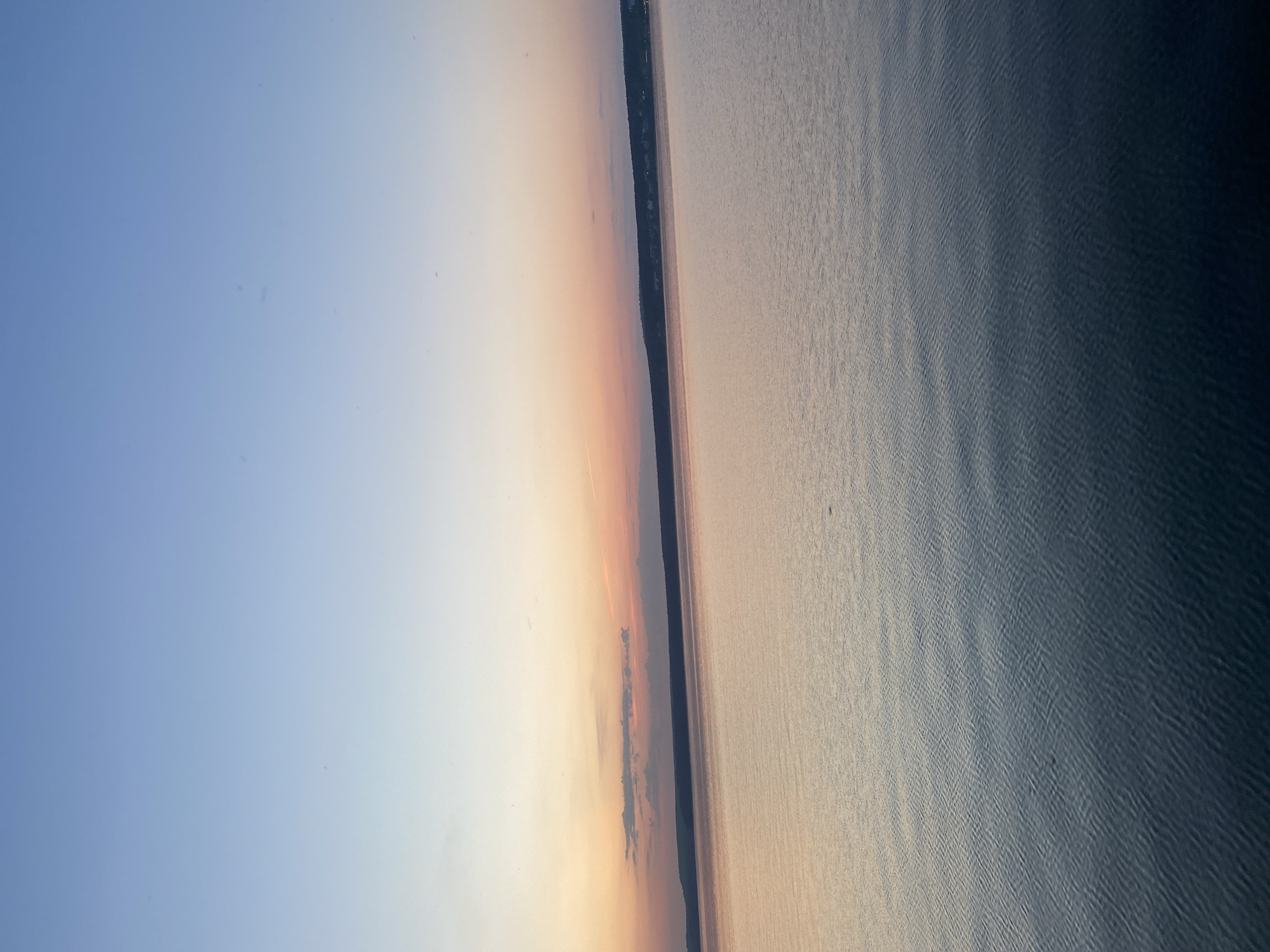 The image depicts a body of water, likely a lake or wide river, under a sky transitioning from light blue to orange-hued clouds near the horizon, suggesting sunrise or sunset. A strip of land with some dark vegetation is visible along the water's edge. Based on the request, it is assumed this is within the city of Votkinsk, Russia. No people, actions or interactions are discernible.