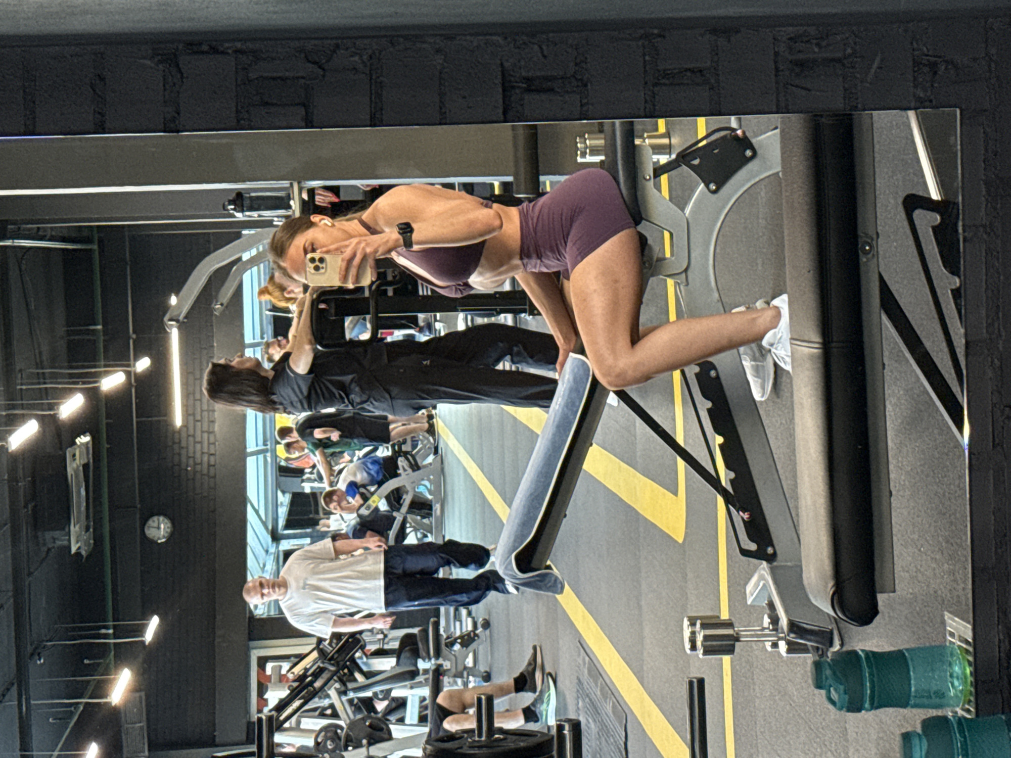 In Saint Petersburg, Russia, a young woman with blonde hair, wearing a purple sports bra and matching shorts, is captured in a mirror selfie at a gym. She is holding a smartphone in her right hand, taking the picture. She is seated on a weight bench, her left hand resting on the bench beside her. A blue towel is draped over the back of the bench. The gym environment is visible, with various weight machines and other gym-goers in the background. A man in a white t-shirt and dark pants is standing in the mid-ground, facing away from the camera. Several other individuals are scattered throughout the gym. The floor is marked with yellow lines. Two green water bottles are visible in the foreground, resting on a rack. The walls of the gym are a mix of dark brick and exposed industrial elements, with fluorescent lighting fixtures on the ceiling.