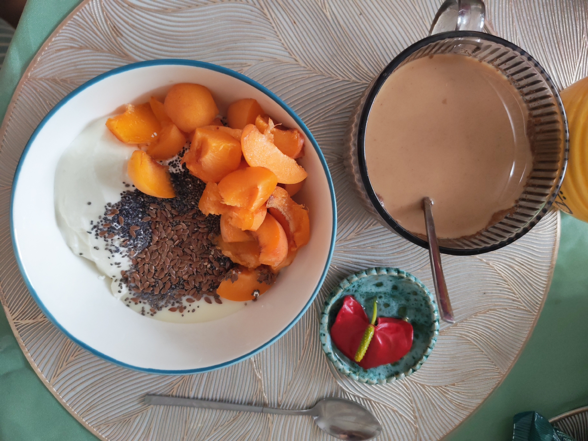 A close-up overhead shot shows a breakfast setting on a table. In the foreground, a white bowl with a blue rim contains yogurt topped with sliced apricots, chia seeds, and flaxseeds. To the right, a glass of a light brown beverage, possibly coffee or tea, is visible. A small decorative dish holds a vibrant red flower with a green stamen. A silver spoon rests horizontally below the bowl, and another spoon is partially submerged in the drink. The table is covered with a textured placemat and a green tablecloth. The background is blurred. The scene appears to be indoors, possibly in Izhevsk, Russia, given the mention of that location. There are no people present in the image.