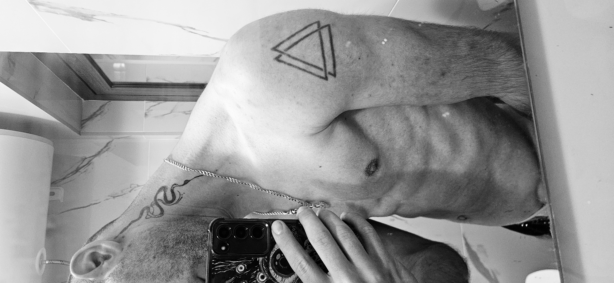 This monochrome mirror selfie, taken in Plovdiv, Bulgaria, captures the upper torso of a person, likely male, rotated horizontally. The individual is taking the photo in what appears to be a bathroom or utility area with light-colored, possibly white, marbled wall tiles.
The main subject displays two prominent tattoos: a geometric design featuring two overlapping triangles on their right shoulder/upper arm, and a stylized snake-like pattern with dot accents on the right side of their neck and upper back. A silver-colored, twisted link chain necklace is visible around their neck. The person's arm and chest show some body hair, and stubble is present on their jawline and around the ear.
The subject is holding a smartphone, a dark-colored Samsung model with three rear camera lenses, encased in a distinctive dark case adorned with multiple stylized eye designs. The hand holding the phone is also visible in the reflection. A white cylindrical object, possibly a water heater or boiler, is partially reflected on the left side of the frame. The image is rich in shadows and reflections, enhancing its depth. No discernible text is visible on the tattoos, phone, or background elements.