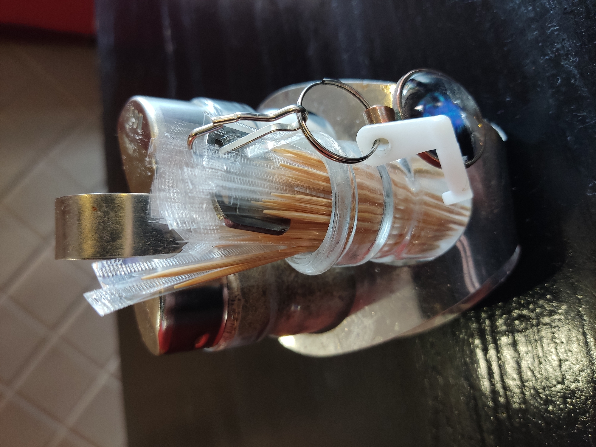 A close-up, eye-level shot shows a condiment holder on a dark, reflective surface, likely a table. The holder is made of metal and contains two glass jars. One jar holds a collection of individually wrapped toothpicks, some of which are made of wood. A metal keychain with a white plastic "L" shaped charm and a round glass charm hangs from the toothpick holder. The second jar appears to contain ground spices, possibly pepper. In the background, a beige tiled wall is visible. The overall scene is well-lit, with some glare on the reflective surface and the metal parts of the holder. The image does not provide enough context to determine its location in Noginsk, Russia.