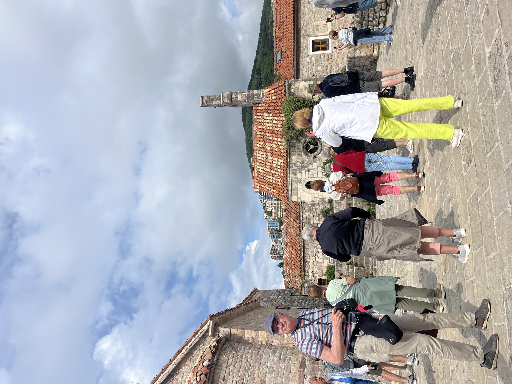 The image shows a group of tourists standing on a stone-paved area, seemingly within the Old Town of Budva, Montenegro. They are looking at or photographing historical buildings. The architecture features terracotta-tiled roofs and stone walls. A tall, partially ruined tower is visible in the background, hinting at the historical significance of the area. The sky is partly cloudy, and mountains can be seen in the distance. The tourists are casually dressed, some carrying cameras, suggesting they are exploring the area.