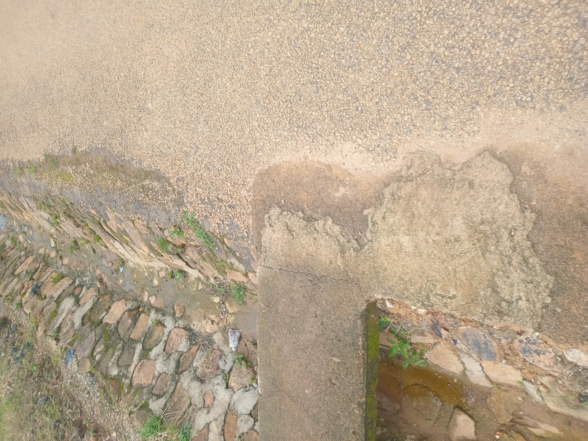 The image depicts a roadside drain filled with water and debris, bordered by a stone-lined embankment. Adjacent to the drain is a paved surface, likely a road or pathway, also showing signs of moisture and discoloration. The scene suggests an outdoor environment in a populated area, possibly within the city of Bukuru, Nigeria, given the contextual information. The overall impression is one of an unmaintained urban drainage system.