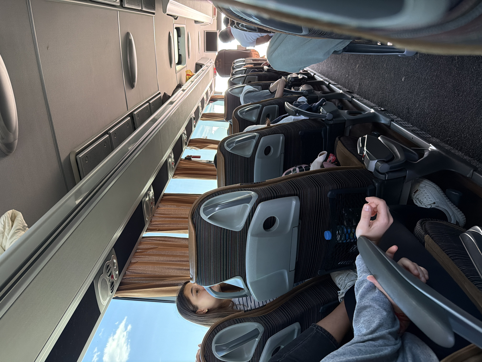 The scene is inside a passenger bus. Rows of seats are visible, most of which are occupied by people. Several passengers are visible, including one wearing a white shirt, and another in a grey sweatshirt. A water bottle is visible in a seat-back pocket. The bus windows show a clear blue sky. Location information (Yazdach, Bulgaria) is not discernible from the image.
