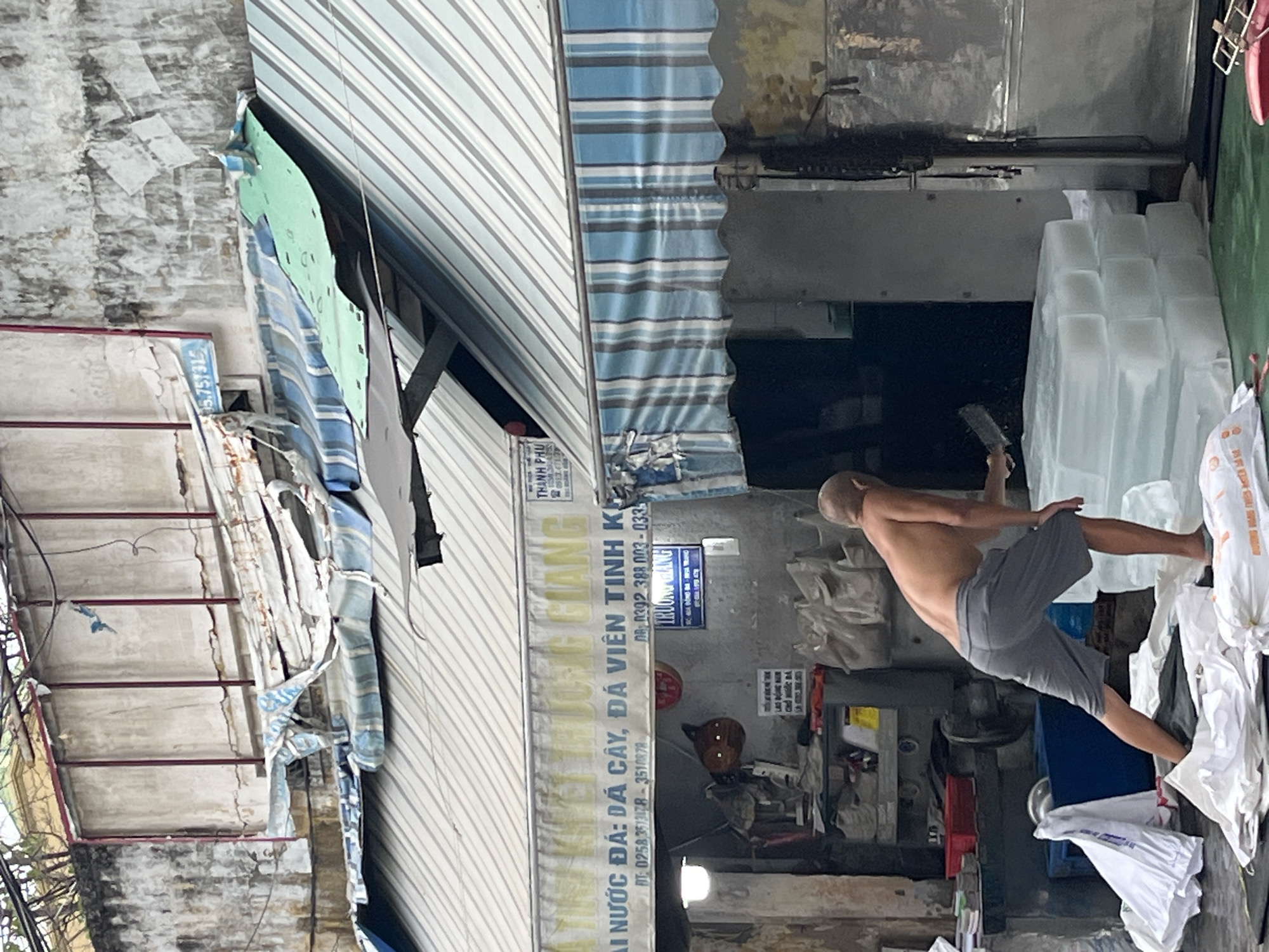 The image depicts an outdoor scene at an ice production facility in Nha Trang, Vietnam, likely a small business. A shirtless man, appearing to be in his late 20s or early 30s, with gray shorts and cropped hair, is actively working. He is bending over and appears to be using a knife or cleaver to cut large blocks of ice, which are stacked nearby. The environment is somewhat cluttered, with various bags, equipment, and awnings visible. The building behind the man shows signs of wear and tear, with peeling paint and damaged awnings. The overall impression is of a working-class establishment focused on the production and sale of ice.