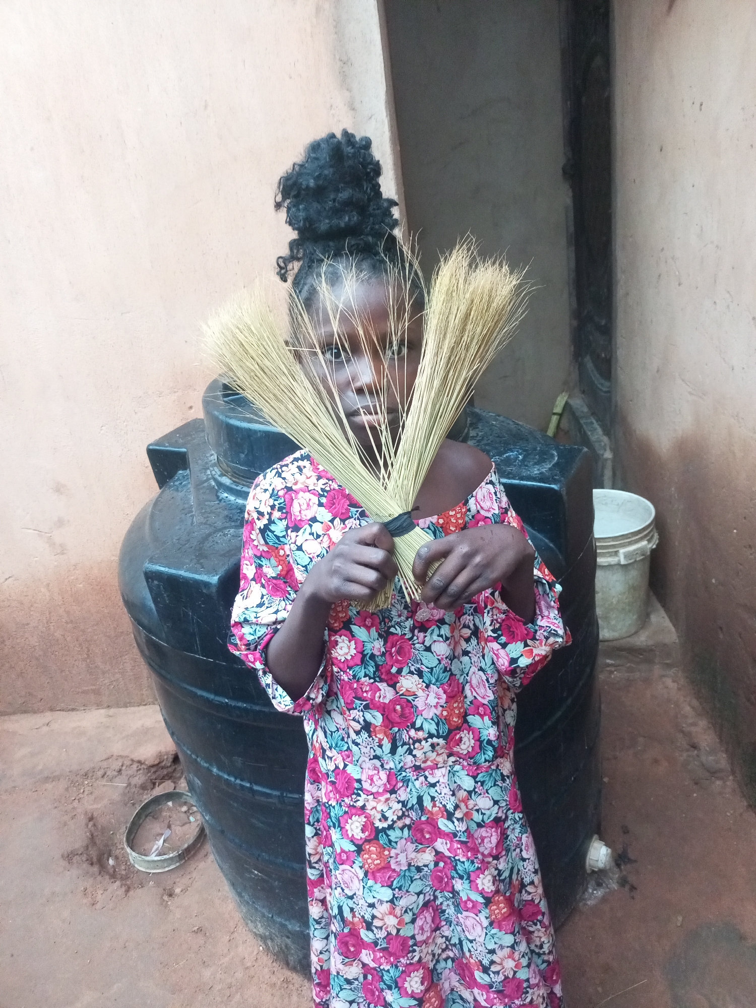 A young Black girl stands in front of a black water tank in what appears to be a rural or semi-urban outdoor setting. She is wearing a brightly colored floral dress with pink, blue, and white flowers. Her dark hair is pulled up in a bun. She is holding a bundle of dried grass, possibly intended for making brooms, in front of her face, obscuring her mouth and nose. The water tank is large and cylindrical, positioned on a dirt or sand-covered ground. Behind the girl and to the right of the water tank, a white bucket is visible. The walls in the background are rough and appear to be made of a light-colored, possibly stucco or mud, material. The overall impression is one of everyday life in a community where such resources and activities are common, consistent with a location in Tempure, Nigeria.