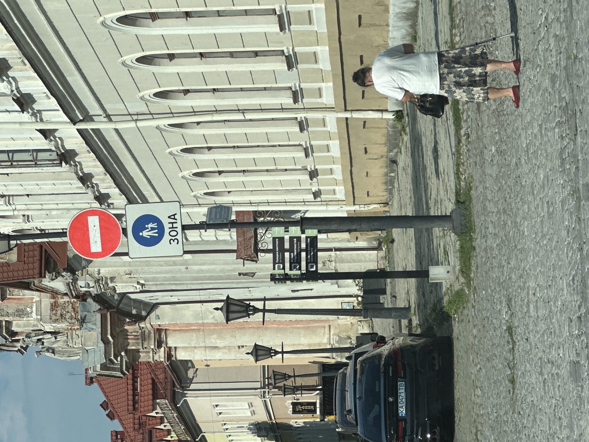 The image shows a street scene in Kamianets-Podilskyi, Ukraine, featuring a cobblestone street lined with historic buildings. In the foreground, a black BMW car with the license plate "KA 0471 TB" is parked. To the right, an elderly woman with her hair in a bun, wearing a white t-shirt and a floral skirt, walks with a cane and carries a dark bag. She is wearing red shoes. Several street lamps are visible along the street, as are various directional signs. One prominent sign features a red circle with a white horizontal bar, indicating "No Entry," and below it, a blue circular sign with a white silhouette of an adult and a child, signifying a pedestrian zone. The buildings exhibit classical architectural elements with arched windows and ornate facades, suggesting a historical district. The sky is clear and blue.