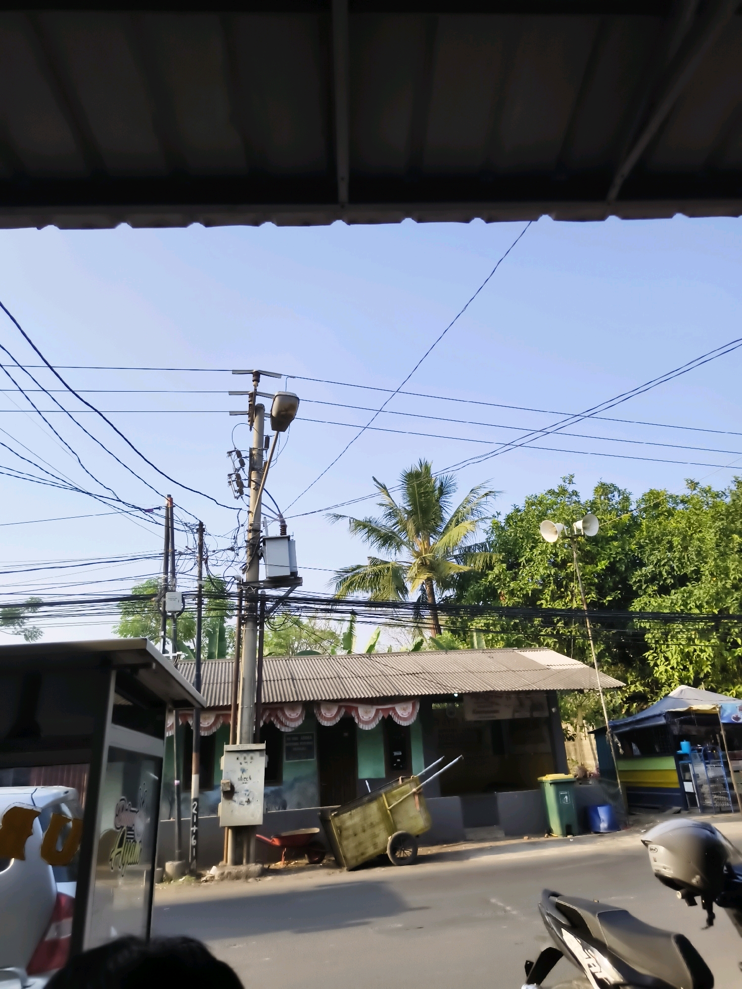 A street scene in Salira, Indonesia is captured on a clear day. The foreground is dominated by the underside of a roofed structure, suggesting a covered walkway or awning. To the left, a portion of a vehicle, possibly a car or bus, is visible, featuring reflective elements and lettering. A person's head is partially visible in the bottom center, indicating the viewpoint of the photograph.
Moving towards the midground, a busy street unfolds with a prominent concrete utility pole at its center, heavily adorned with a tangled network of electrical wires. A transformer box is mounted on the pole, along with a street lamp and various electrical components. Adjacent to the utility pole, a small building with a corrugated metal roof and decorative awnings is situated. In front of this building, a yellow handcart with two wheels is parked. Further to the right, another stall or kiosk with a blue and yellow striped awning is present, with shelves visible behind the counter. A green trash bin stands next to the kiosk.
The background is filled with lush green trees, including a palm tree, suggesting a tropical environment. The sky is a clear, pale blue, indicating fair weather. A motorcycle is parked in the lower right corner of the frame, with its helmet resting on the seat. The scene appears to be a typical urban street in Indonesia, characterized by utility infrastructure, small businesses, and natural vegetation.
