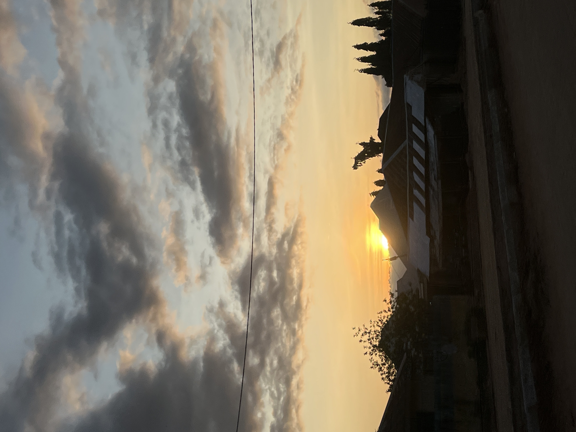 This image depicts a rural or semi-urban landscape in Howayi, Nigeria, at either sunrise or sunset. The upper sky features a mix of dark grey and white clouds against a light blue backdrop, with orange and pink hues appearing lower down due to the sun's angle. A single, dark power line stretches horizontally across the mid-sky. The sun, a bright orange-yellow orb, is partially visible low on the horizon, casting a strong warm glow that illuminates the lower clouds and distant atmosphere. The foreground and midground are rendered in dark silhouette, showing multiple low-rise residential structures with distinct rooflines, some appearing to be made of corrugated metal, with one roof reflecting a sliver of sunlight on the right. Silhouetted trees are visible, including a sparse deciduous tree on the left and a cluster of tall, conical trees on the far right. The very bottom of the image shows a dark, unpaved road or path with a visible curb. No people, animals, or explicit actions are discernible within the frame.