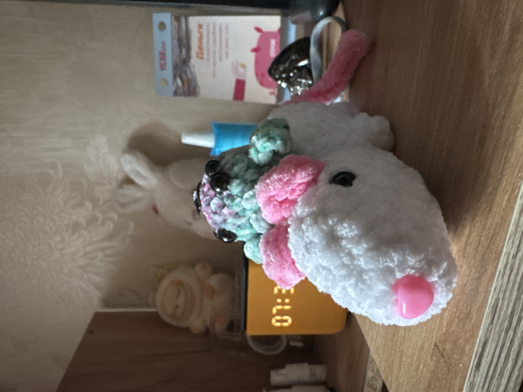 A stuffed unicorn toy with white, pink, and pastel green yarn, and black button eyes, rests on a wooden surface. The unicorn has pink ear-like accents on its head and a pink tail. A pink heart-shaped nose is visible on its snout. Behind the unicorn, an orange digital clock displays "07:3". To the left, a small, light-colored figurine sits on a shelf. In the background, a white stuffed bunny is partially visible, along with a blue dispenser and a calendar featuring an illustration of pink pigs. The scene appears to be an indoor setting, possibly a bedroom or living area. There is no discernible connection to Nizhny Novgorod, Russia.