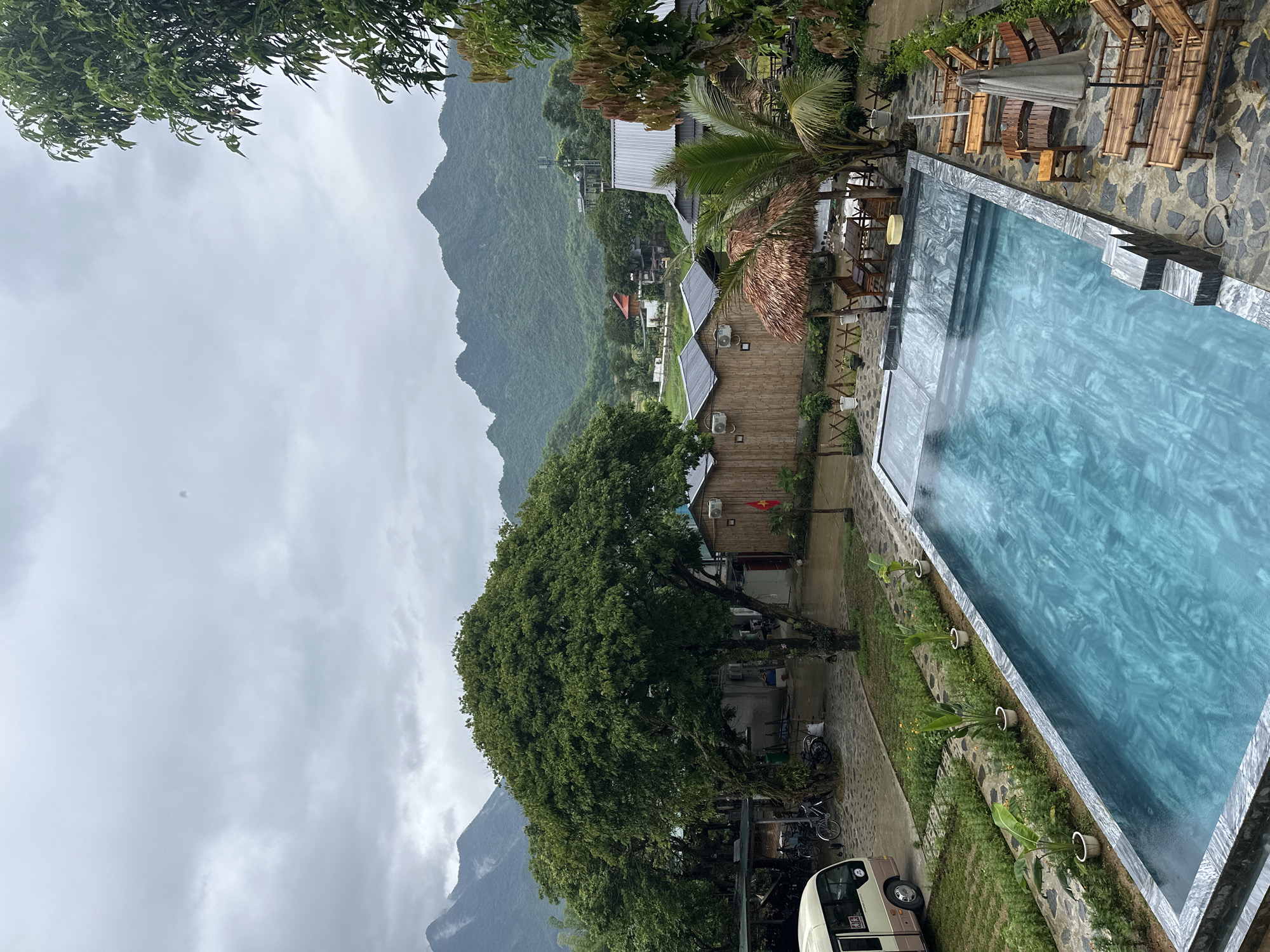The image shows a view of a swimming pool and resort area with a mountainous backdrop in Xóm Khán, Vietnam. The sky is overcast with gray clouds. In the foreground, a rectangular swimming pool with blue, marbled tiling is visible. Several wooden lounge chairs with umbrellas are arranged around the pool. Behind the pool, there are thatched-roof structures, palm trees, and a few bamboo bungalows with air conditioning units. To the left, a small white and beige bus is parked, and bicycles are leaning against a covered area. A large, leafy green tree stands prominently near the center of the image. In the background, rolling green mountains are covered in lush vegetation. Small houses and other buildings are scattered among the trees.