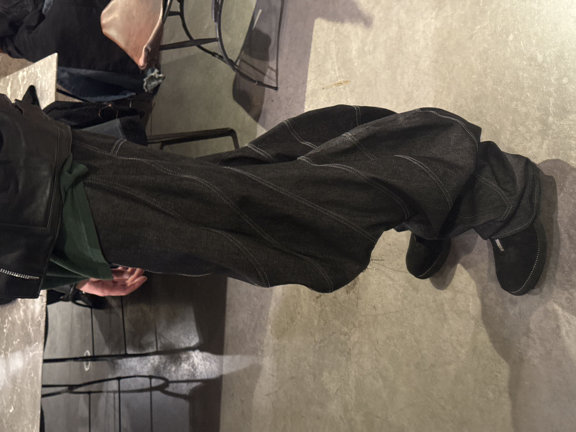 A person is standing indoors in what appears to be a restaurant or café. They are wearing dark, baggy denim pants and black shoes. They are also wearing a black leather jacket and a green shirt is visible underneath. The floor is concrete and there are chairs and tables in the background, with one table having a marble top. The location cannot be definitively identified as Paris, France from the given image.