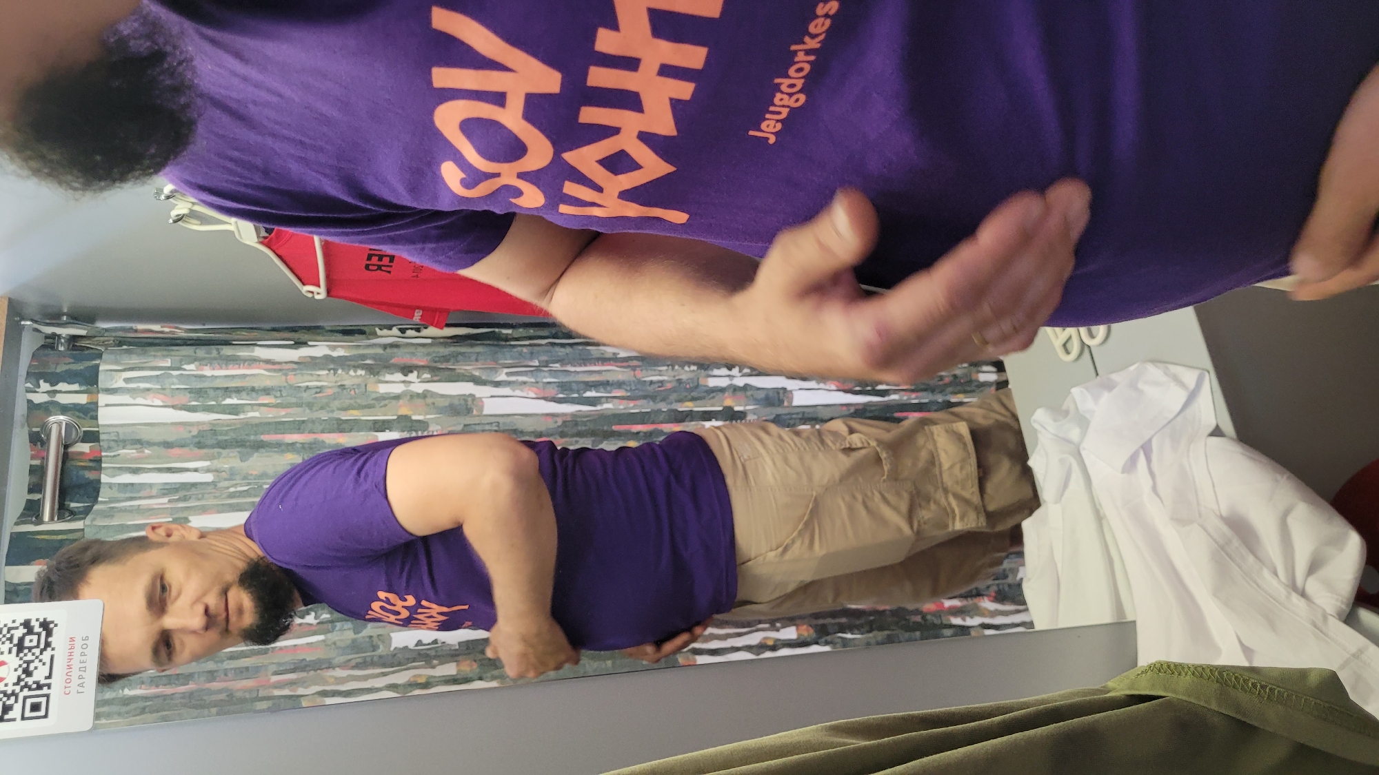 A mirrored image shows a man in a changing room. He is wearing a purple t-shirt with orange text and khaki pants. The text on the shirt appears to read "SOV KAUT Jeugdorkes". The changing room has a shower curtain with a patterned design. There is a white shirt hanging on a hanger in the background. The mirror has a QR code sticker in the lower left corner. The scene is located in an unknown city.
