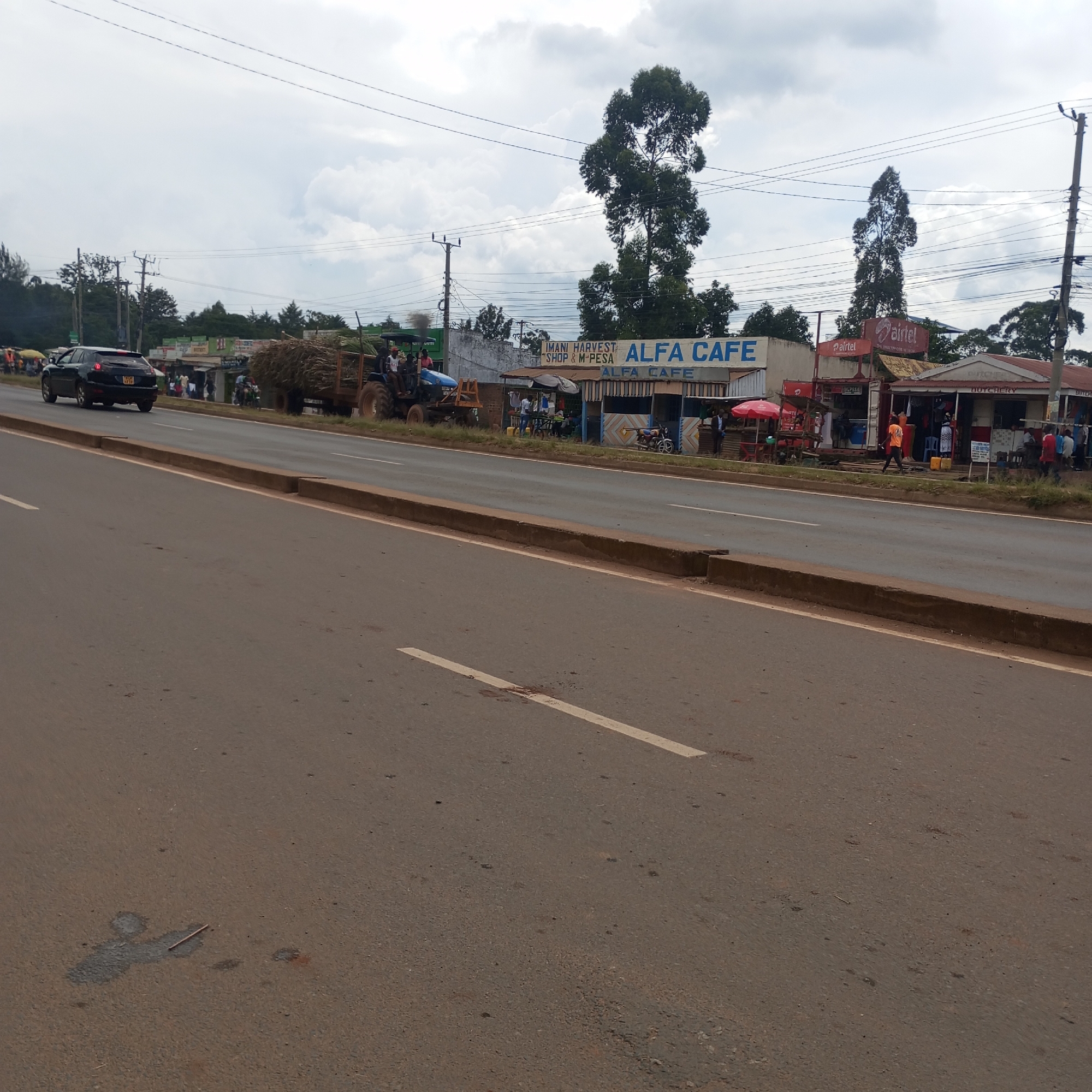 The image depicts a street scene in Sheywe ward, Kenya. A car is traveling on a two-lane road with a concrete median. On the right side of the road are several small businesses, including Alfa Cafe and a butcher shop. There are people walking and standing in front of the shops. A tractor carrying a load of material is also visible on the right side of the road. Overhead power lines are strung across the scene. The sky is cloudy.