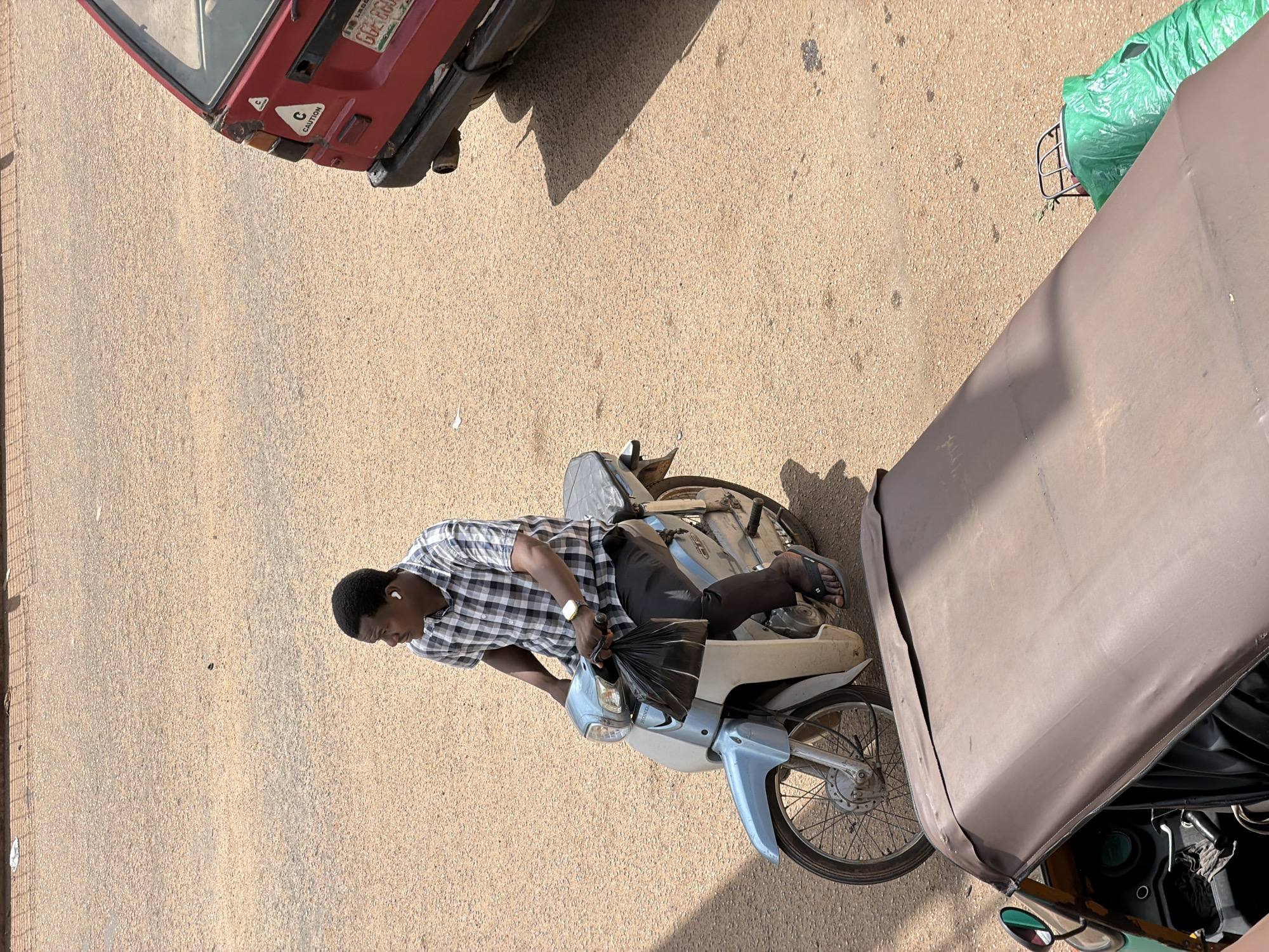 In Bukuru, Nigeria, a man wearing a checkered shirt and black pants rides a light blue and white motorcycle. He has on black flip-flops and is wearing white earbuds and a wristwatch. He holds a black plastic bag in his left hand. In the background, a red car is parked, and a portion of a green and brown auto-rickshaw is visible in the foreground. The ground is a sandy, asphalt surface, typical of Nigerian streets.