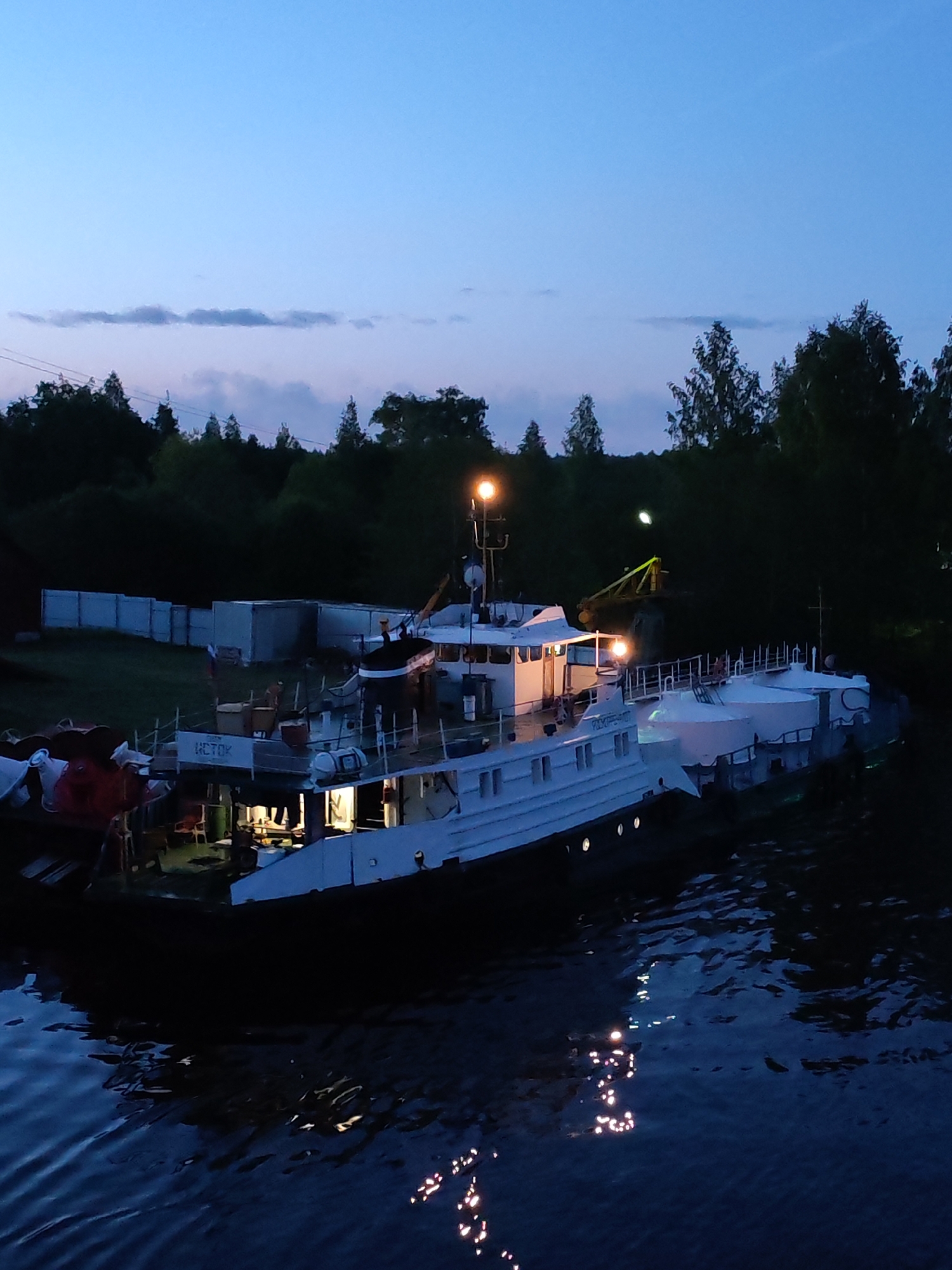 A white ship with lights on, labeled "ICTOK," is docked on a body of water, likely a river or canal, at night. A white fence and trees are visible in the background. The location is likely Annensky Most, Russia, based on the ship's Russian labeling. The water reflects the ship's lights.