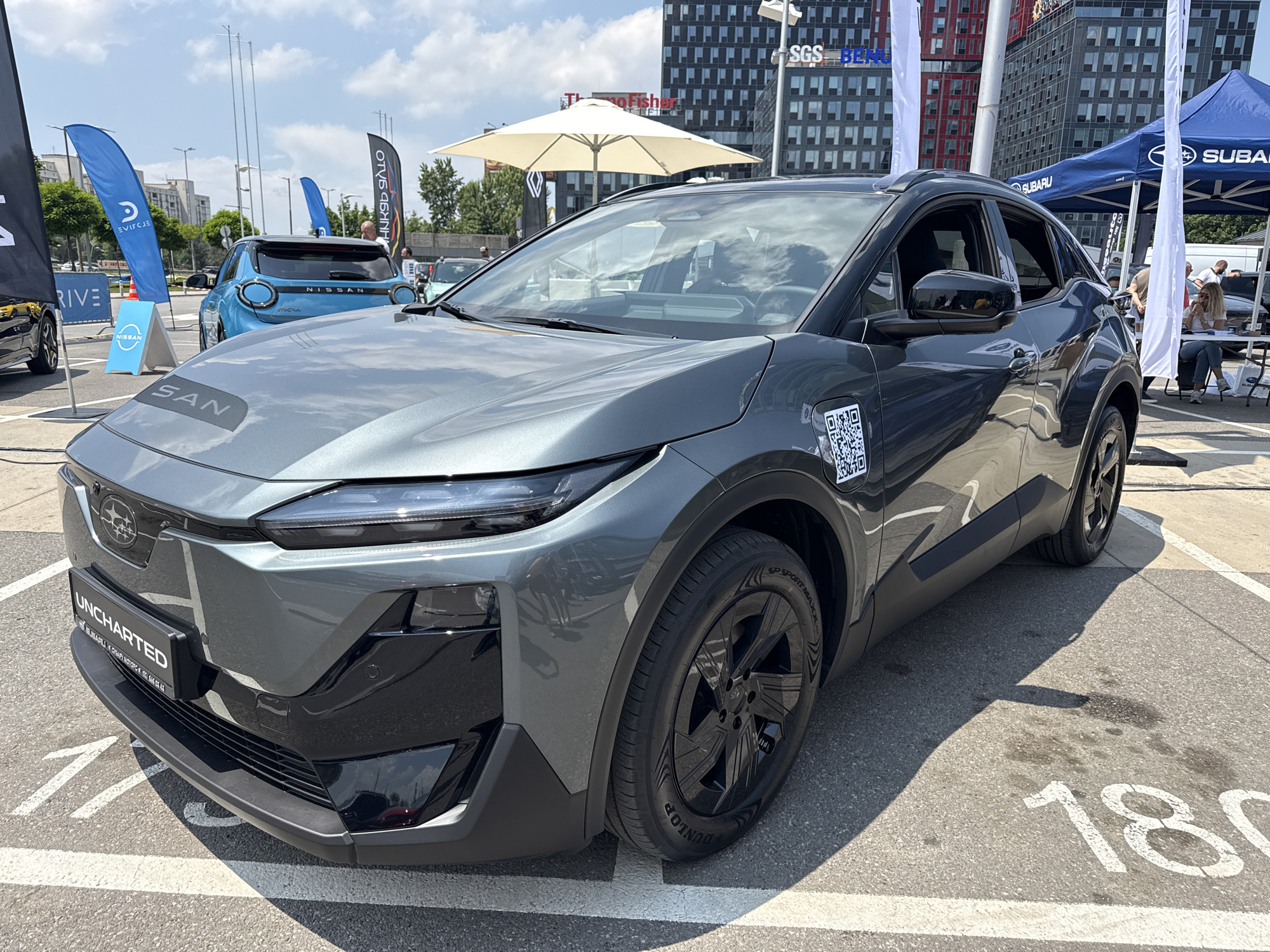 This image captures an outdoor car exhibition or promotional event on a sunny day, likely in the Sofia Tech Park area of Sofia, Bulgaria.
In the foreground, a dark grey metallic Subaru crossover SUV is prominently displayed, parked on asphalt with white painted parking lines, including a partially visible "180". The vehicle features a Subaru badge on the front, sleek LED headlights, a black lower bumper and grille section, and black multi-spoke wheels fitted with Dunlop SP Sport Maxx tires. A black rectangular badge on the lower front grille reads "UNCHARTED", and beneath it, smaller text indicates "SUBARU DREAMS MOTORS" along with a phone number. A white QR code sticker is affixed to the front passenger-side fender.
In the mid-ground, to the left of the Subaru, a light blue Nissan crossover SUV is visible from the rear, featuring circular rear light designs and "NISSAN" branding. Several promotional flags are present: a black flag partially visible on the far left reading "DRIVE", a blue feather flag with a white logo and reversed text, and a blue A-frame sign displaying the Nissan logo.
To the right of the Subaru, a blue tent with the "SUBARU" logo is set up, under which a blonde woman in a white top and jeans is seated at a white table, looking at a phone or device. Other indistinct figures are visible around the tent. A large white patio umbrella provides shade behind the Subaru. Various other flags and banners are scattered throughout the background, including one black feather flag with reversed text.
The background consists of modern, multi-story commercial buildings with glass and dark facades. Identifiable corporate logos include "SGS" and "BERH" on one building, and "ThermoFisher" on another, which are consistent with businesses located within Sofia Tech Park. The sky is partially cloudy with bright sunlight casting distinct shadows. Trees are sparsely visible between the foreground and background structures.