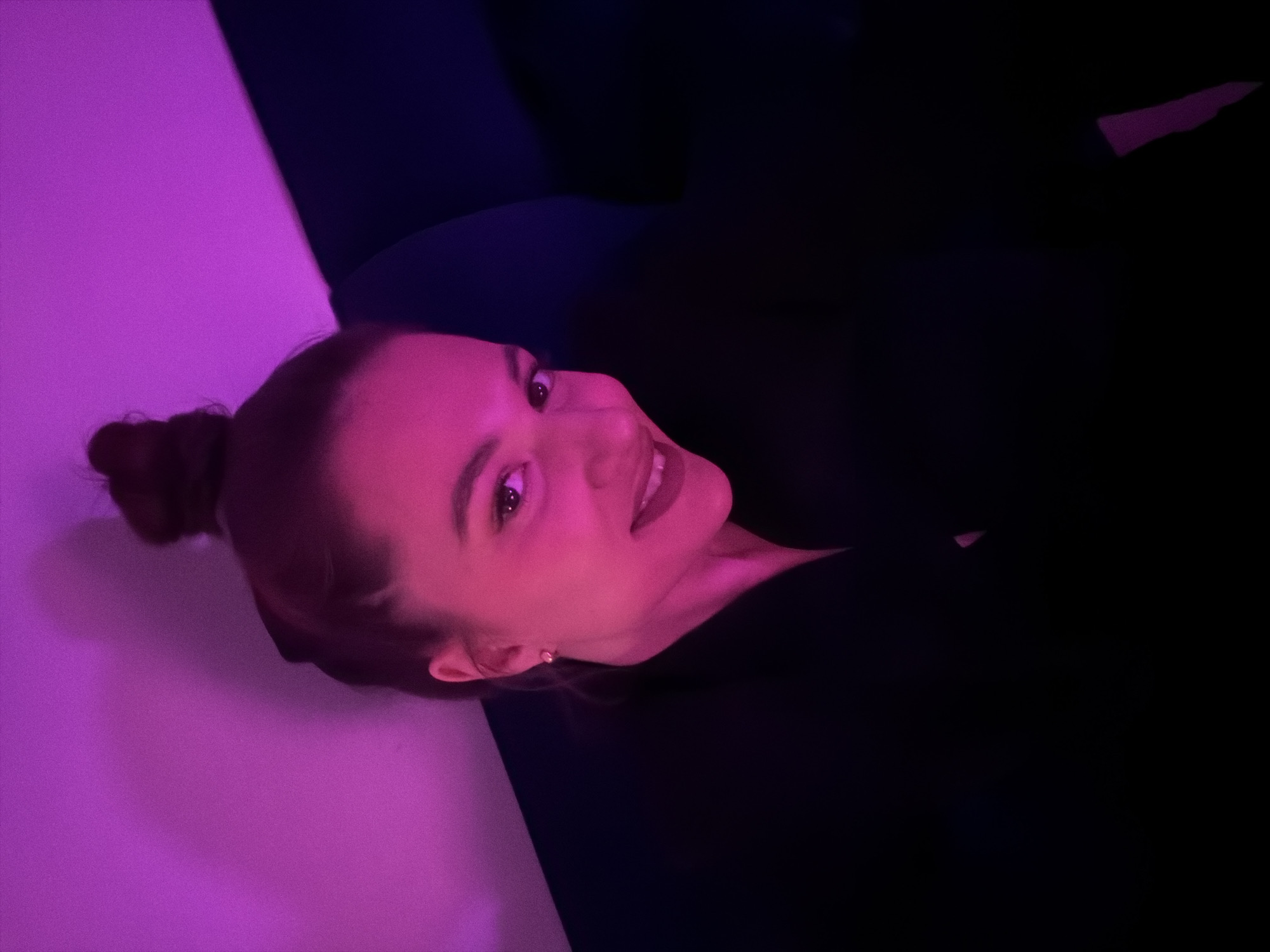 A young woman is shown in a low-angle shot, likely in an indoor setting. She has her hair in a bun and is wearing a dark-colored top. The lighting in the scene is heavily tinted with purple and deep blue hues. A white surface, possibly a wall, is illuminated in the bottom left corner by purple light. The backdrop is mostly dark, obscuring details of the environment. There's no specific indication that this image was taken in Belgrade, Serbia.