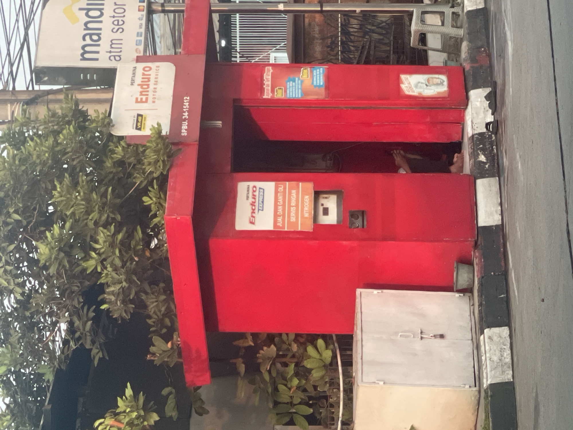 The image shows a red kiosk with a sign that reads "PERTAMINA Enduro MOTOR SERVICE" and "SPBU. 34-15412". On the right side of the kiosk is a sign that says "Mandiri ATM Setor". There is a person sitting inside the kiosk. The kiosk is located on a street with a curb and a sidewalk. There are trees and plants around the kiosk. The scene appears to be in South Tangerang, Indonesia.