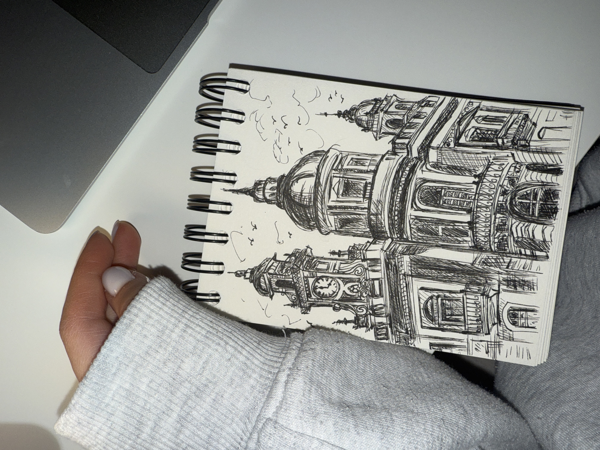 An indoor shot captures a person's hand holding open a spiral-bound sketchbook, revealing a detailed architectural drawing. The hand, seen from the side, wears a light grey long-sleeved top, possibly a sweatshirt. The sketchbook is open to a page featuring a black ink drawing of a grand, ornate building with multiple towers and domes.
The drawing depicts a large structure characterized by intricate details, including arched windows, balustraded balconies, and decorative facades. One prominent tower on the left features a visible clock face, with hands indicating approximately ten past ten or ten to eleven. Several smaller, abstract shapes, resembling birds or clouds, are sketched in the white space above the building.
Given the context of Sofia, Bulgaria, the architecture strongly suggests the **Central Military Club** or a similar historic landmark in the city. The drawing style is detailed, with cross-hatching and fine lines used to create texture and depth.
Part of a dark grey laptop is visible in the upper left corner of the frame, suggesting a workspace setting. The background is a clean, light-colored surface, likely a desk. There is no visible text in the image itself, nor are specific emotions discernible. The lighting is even, suggesting an indoor setting without strong natural light sources.