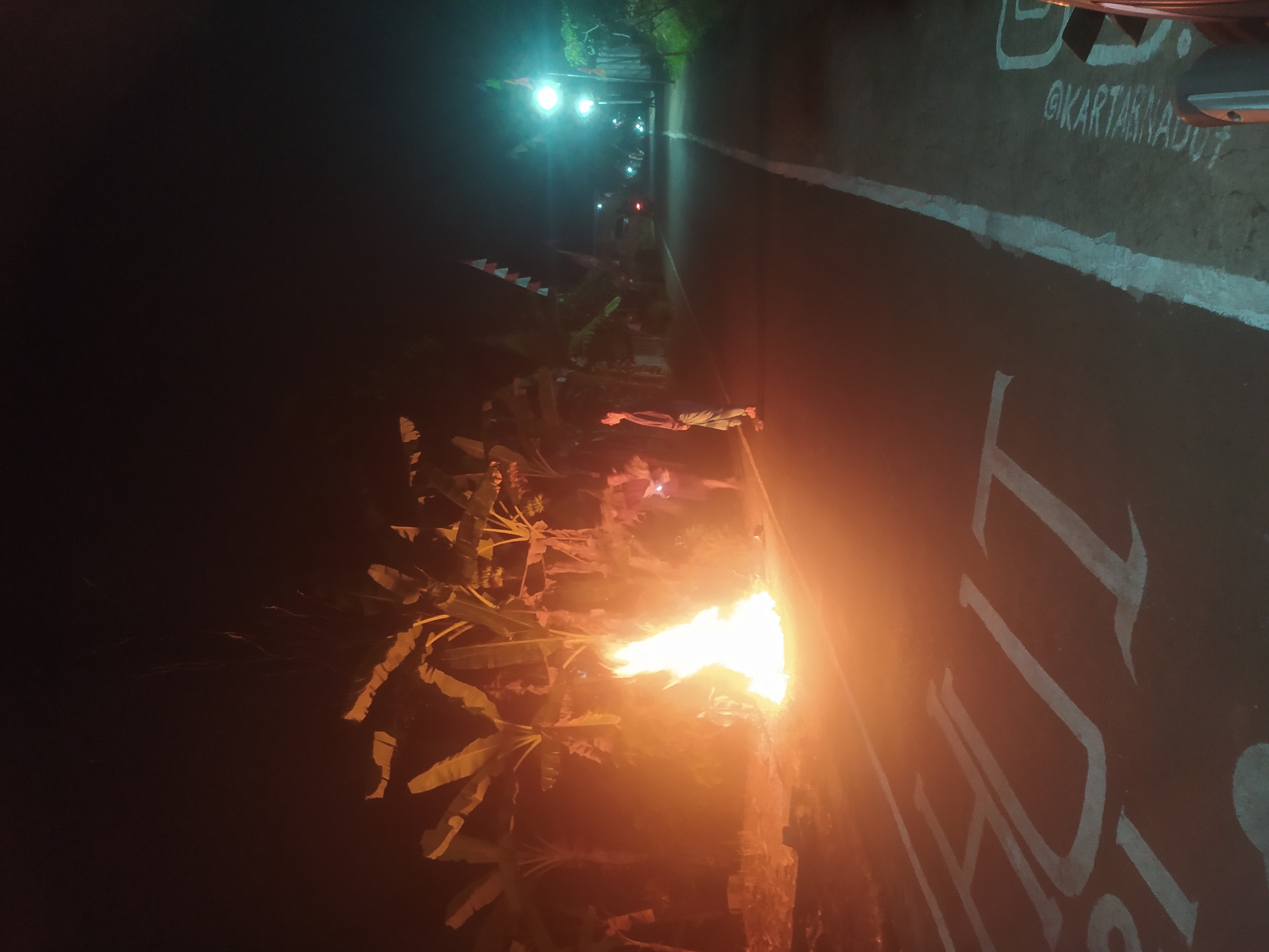 In Lopait, Indonesia, at night, a bonfire burns brightly on the side of a dark asphalt road. The flames are tall and cast an orange glow, illuminating the surrounding banana trees and the road surface. Two individuals are present. One person stands near the bonfire, their form partially obscured by the flames. Another person, wearing light-colored pants and a dark top, stands further down the road, facing away from the bonfire. The road has white painted markings, including what appears to be the word "HUT" painted in large white letters on the left side and "@KARTARINAJUUT" in smaller white lettering on the right. Streetlights illuminate the road in the distance, and flags, possibly for a celebration, are strung along the roadside. The scene suggests a nighttime gathering or event.