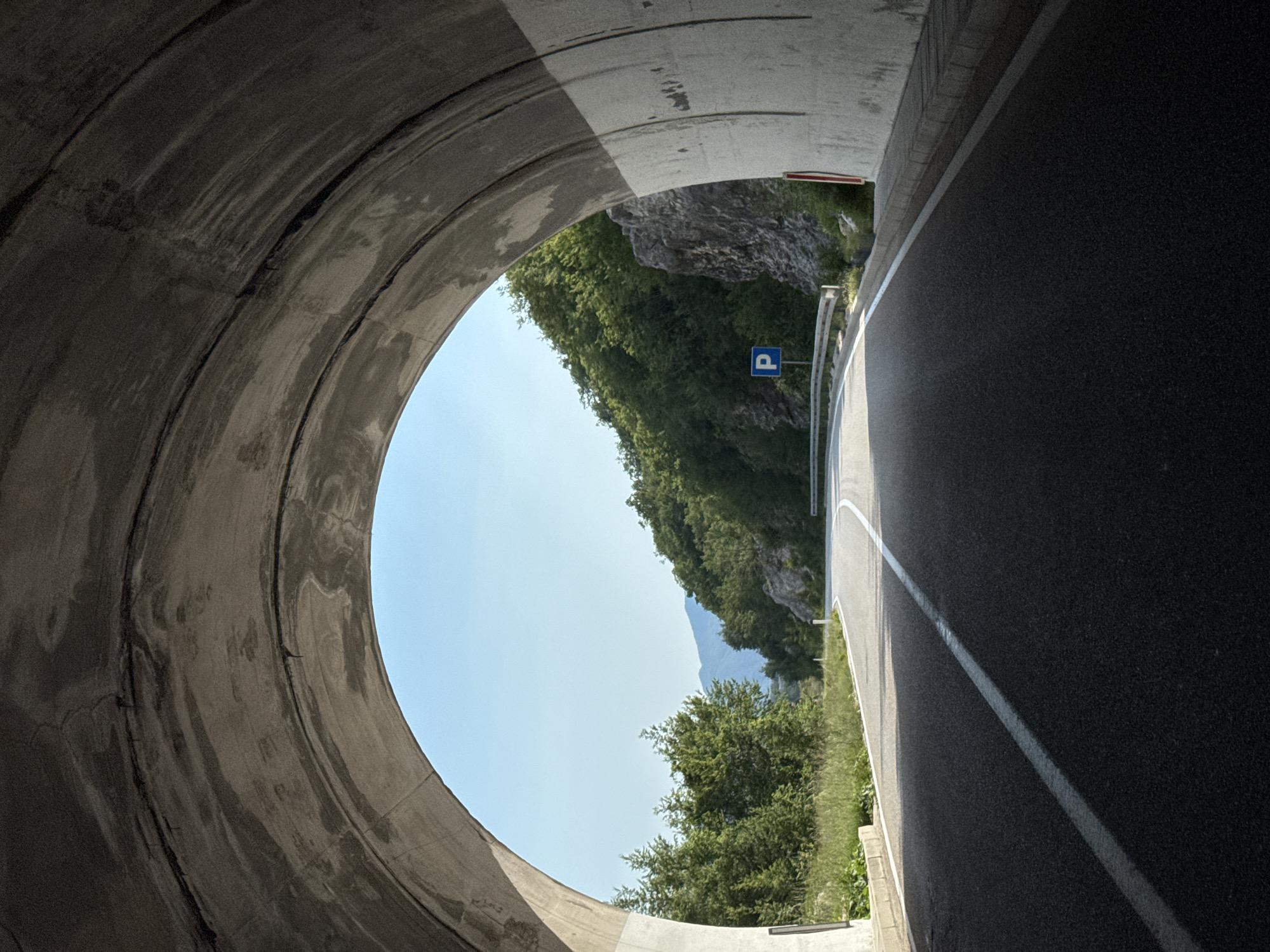 The media file shows a view from inside a concrete tunnel, looking out towards a bright, clear sky and a lush green mountainside. The tunnel opening is circular and imposing, with rough concrete textures and visible lines of construction. On the right side of the frame, a paved road curves away, marked with a white line. Beyond the road, the mountainside is covered in dense green trees, with a rocky cliff face visible higher up. A blue parking sign with a white "P" is mounted on a pole beside the road, indicating a possible resting or viewing area. The time of day appears to be midday, with strong sunlight casting shadows. The weather is clear and fair. The overall scene suggests a scenic route through mountainous terrain, likely in a rural or natural setting. The image evokes a sense of transition, moving from the dark, enclosed space of the tunnel to the open expanse of nature. There is no visible text other than the letter "P" on the parking sign. The location is identified as Dragalj, Montenegro.