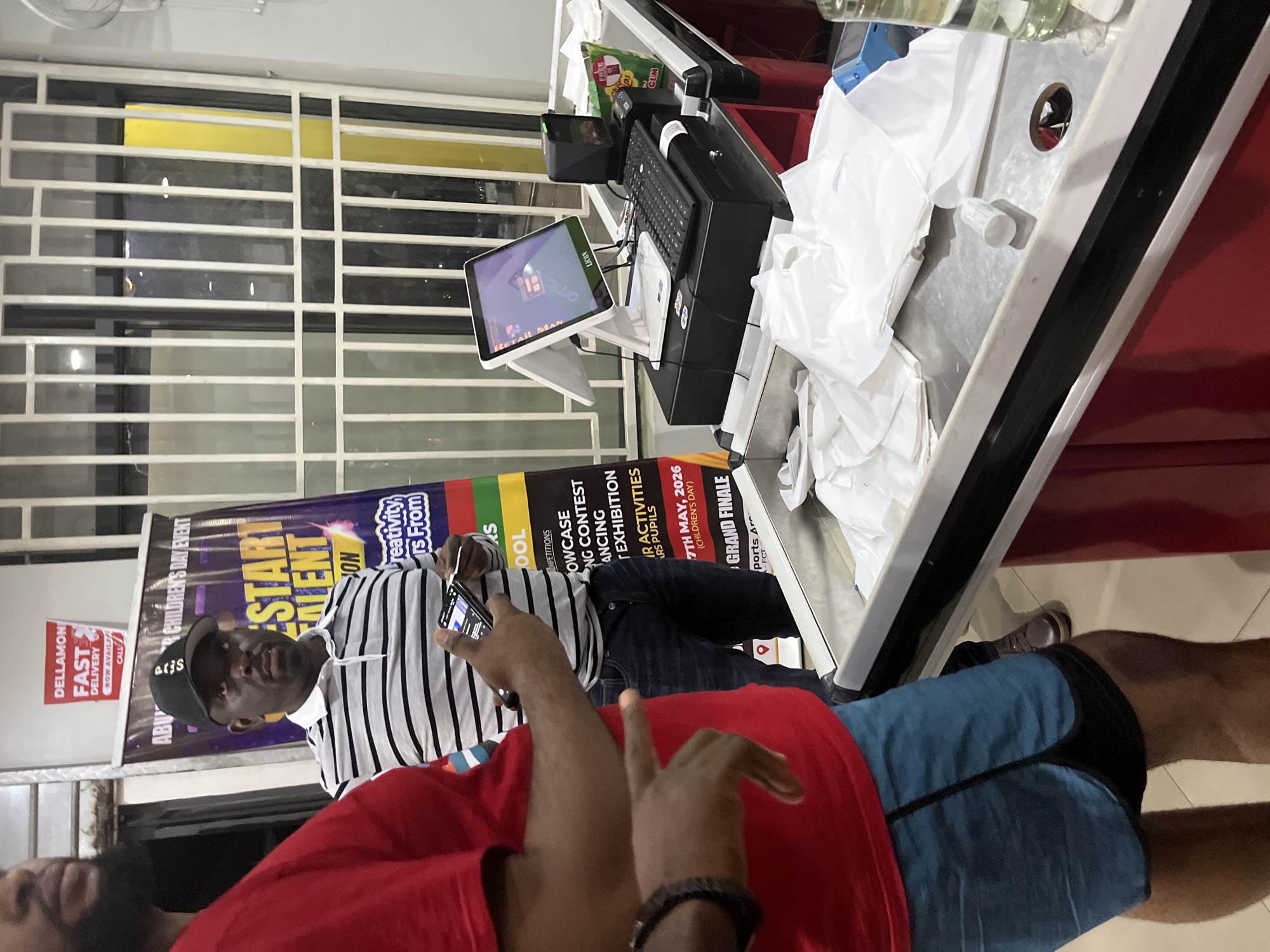 Here is a description of the image:
**Scene:** An indoor space, likely a commercial establishment in Abuja, Nigeria.
**People:** At least three individuals are partially visible. One man is wearing a black baseball cap with the word "Aris" on it, along with a striped shirt. He is holding a mobile phone. Another man is wearing a red shirt, and part of his face is visible. A third individual is partially visible with blue shorts and brown shoes.
**Objects:** The scene is dominated by a point-of-sale (POS) system, including a computer screen, a keyboard, and likely a cash register. There are shopping bags in the bagging area. A poster advertising "Abuja Children's Day Event" is behind the man with the striped shirt. The poster indicates the date "7th May, 2026." There is also text advertising Dellamon Fast Delivery.
**Actions/Interactions:** The man with the striped shirt appears to be using his mobile phone. The other individuals are in close proximity.
**Environment:** The background includes a grated window. The POS system is positioned on a counter, suggesting a retail or customer service environment. The lighting suggests an indoor setting.
