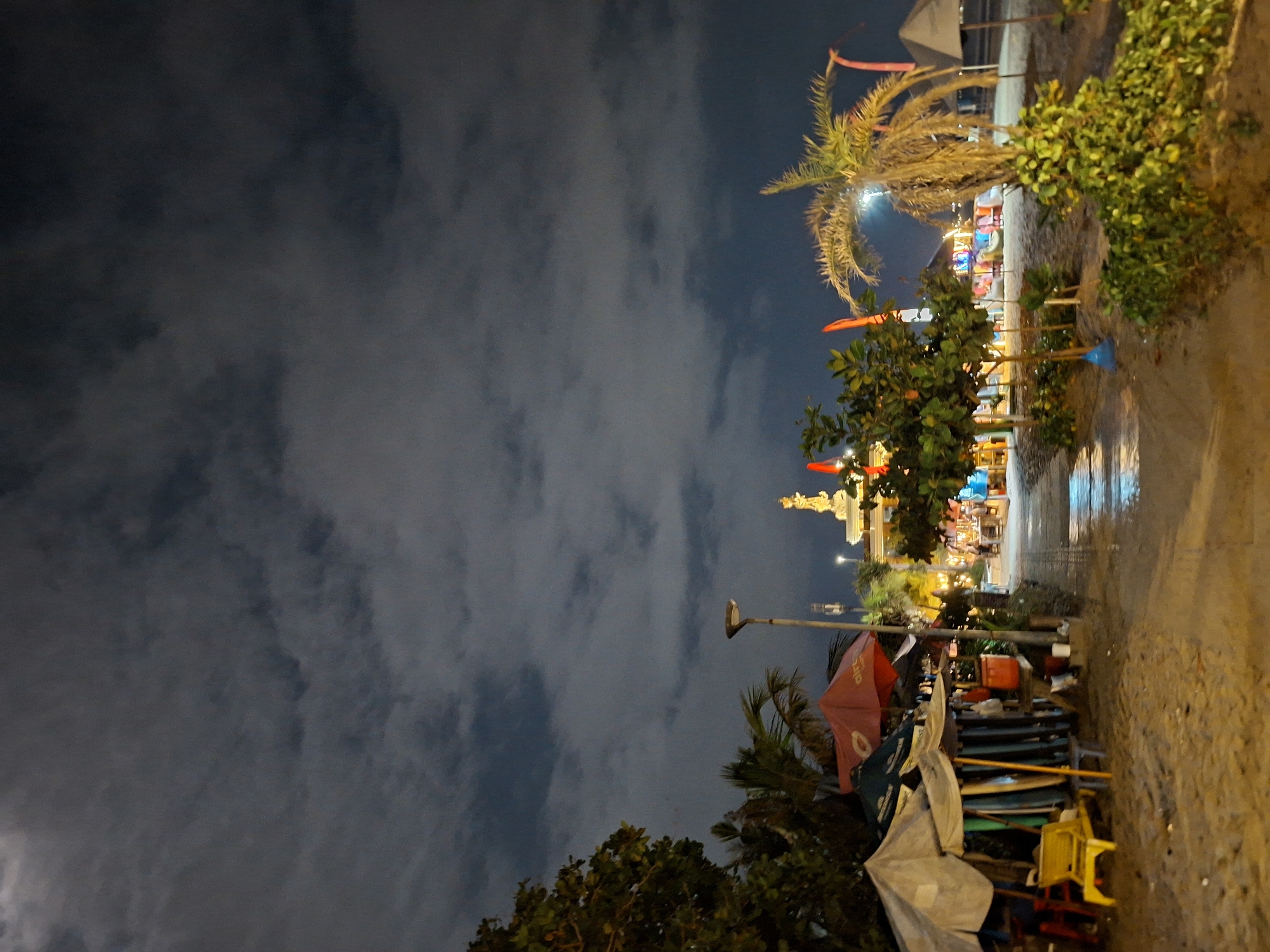 A nighttime scene on Kuta Beach, Bali, Indonesia. The sky is dark with heavy clouds. In the foreground, a sandy beach stretches towards a pathway. To the left, several beach umbrellas are set up, with surfboards leaning against them. A yellow beach chair is visible. Further down the path, there are more umbrellas, trees, and outdoor seating areas with people. A statue can be seen in the distance, illuminated by lights. The lighting suggests artificial illumination from the establishments along the beach. The overall atmosphere is calm and leisurely.