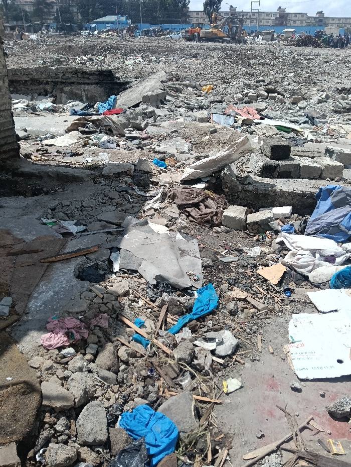 This image depicts a vast demolition site in Nairobi, Kenya, consistent with the Kariobangi demolitions. The scene is an expansive, uneven landscape of pulverized concrete, broken masonry, twisted rebar, splintered wood, and dark earth, indicative of recently destroyed structures.
In the immediate foreground, assorted debris includes large concrete chunks, smaller stones, wood fragments, and scattered personal effects. Multiple bundles of fabric are visible: a light pink/red bundle near the lower left, a dark brown bundle centrally, and several bright blue bundles distributed across the mid-foreground. A black plastic bag is present near one blue bundle, and a white cardboard box with faint text lies on the right.
The midground extends the field of rubble, showing larger concrete slabs and an excavated depression or foundation pit. In the background, a yellow excavator is visible, actively involved in clearing or demolition work. Several individuals are present near the excavator and further in the distance, consistent with workers or displaced persons observing the site. Distant multi-story buildings, some featuring light blue facades or roofs, delineate the far horizon under a bright, partially clouded sky.