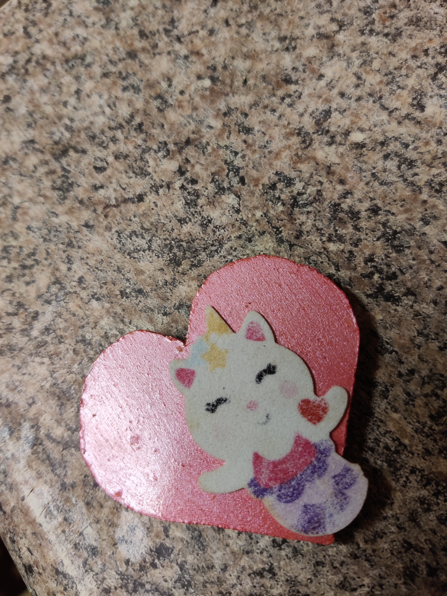 A pink heart-shaped object with a white cartoon unicorn sticker is placed on a speckled countertop. The unicorn has a yellow horn, black eyes, pink cheeks, and a red heart on its cheek. It is wearing a purple and pink patterned scarf. The countertop has a textured, granite-like appearance with brown, black, and tan speckles. The lighting in the image is slightly dim, casting a shadow on the right side of the heart. The photograph was taken from a top-down perspective. This image does not contain any information about a specific location in Lower Paxton Township, United States of America.