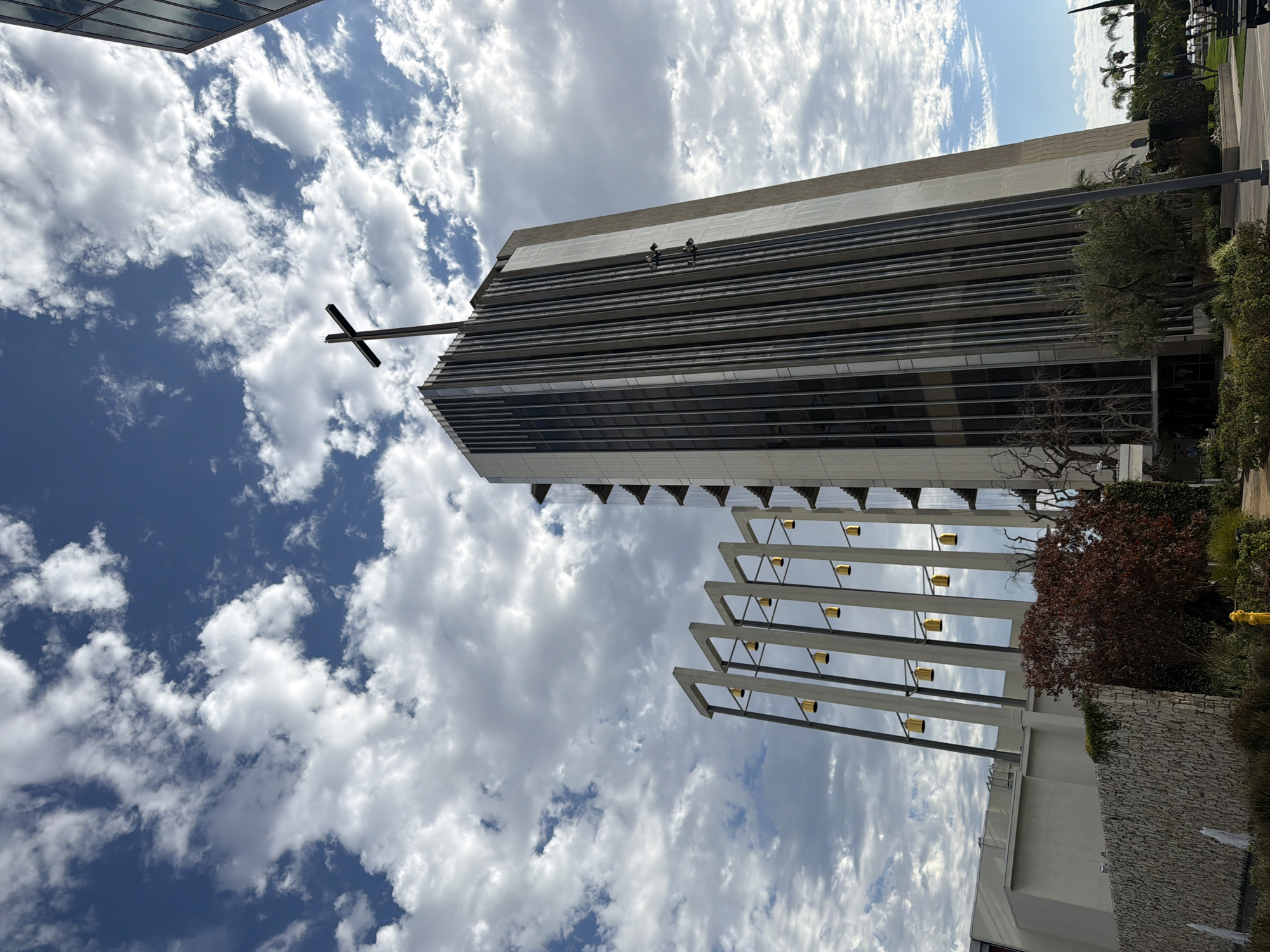 This is a low-angle, full exterior, eye-level shot of Christ Cathedral in Garden Grove, California, United States of America. The church, formerly the Crystal Cathedral, features a tall, modern tower with a cross at its apex. The tower is flanked by a structure with vertical lines and a series of golden bells suspended from angled supports. The building is made of concrete and glass. In the foreground, a stone wall runs horizontally across the frame, with a small fountain and some vegetation. The sky is blue with scattered white clouds. A person is visible on a distant bench.