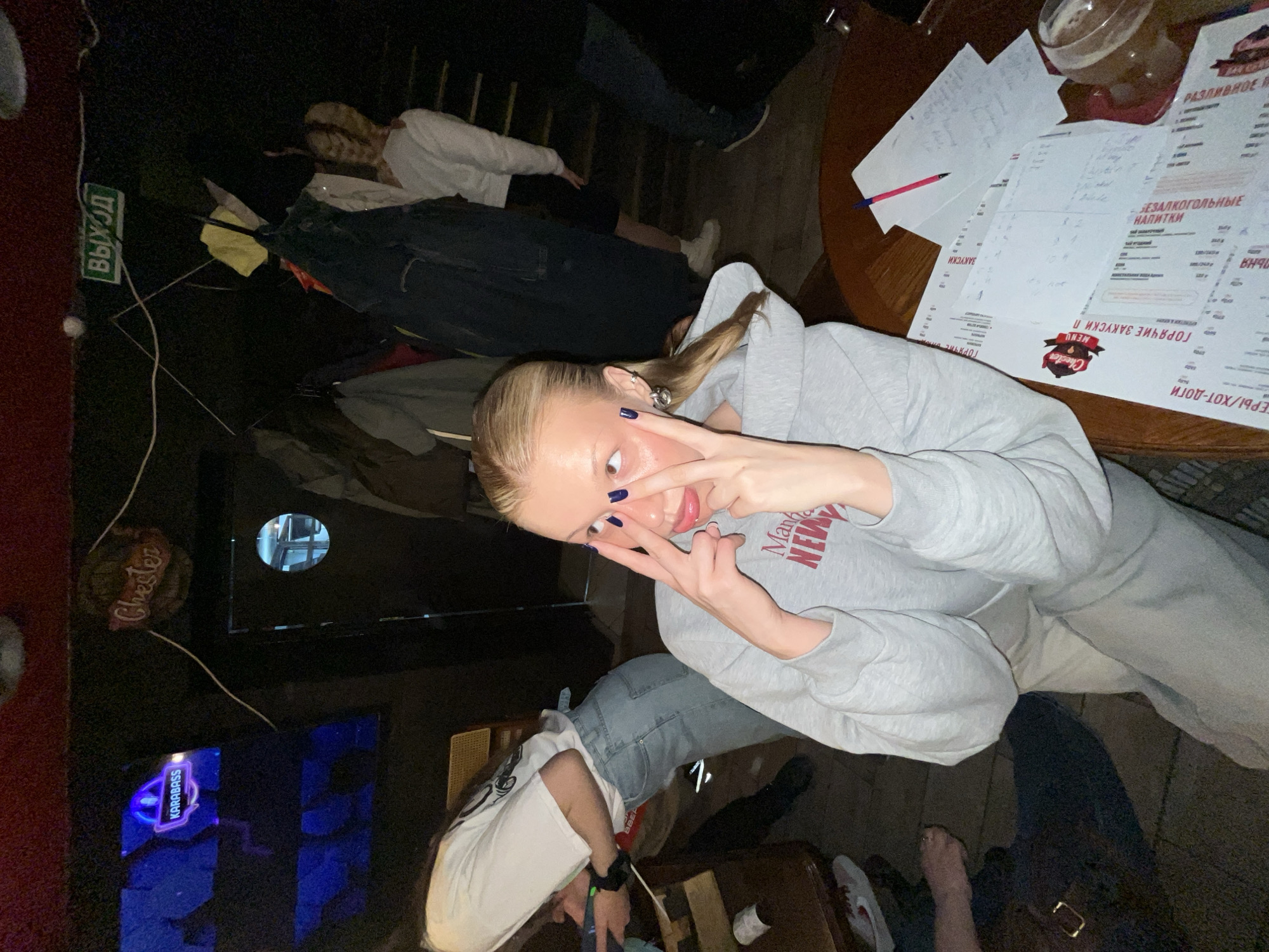 A woman with blonde hair and dark blue nail polish makes a "peace" sign with her fingers near her face. She wears a grey hooded sweatshirt with the word "Manhattan" on it in red. She is sitting at a wooden table in a dimly lit place, possibly a restaurant or bar in Chaikovsky, Russia. Papers and a menu are visible on the table. The "Chester" menu suggests the location of the bar/restaurant. There are other people in the background, and signage indicating an exit. A Karabas bar sign is seen in the distance.