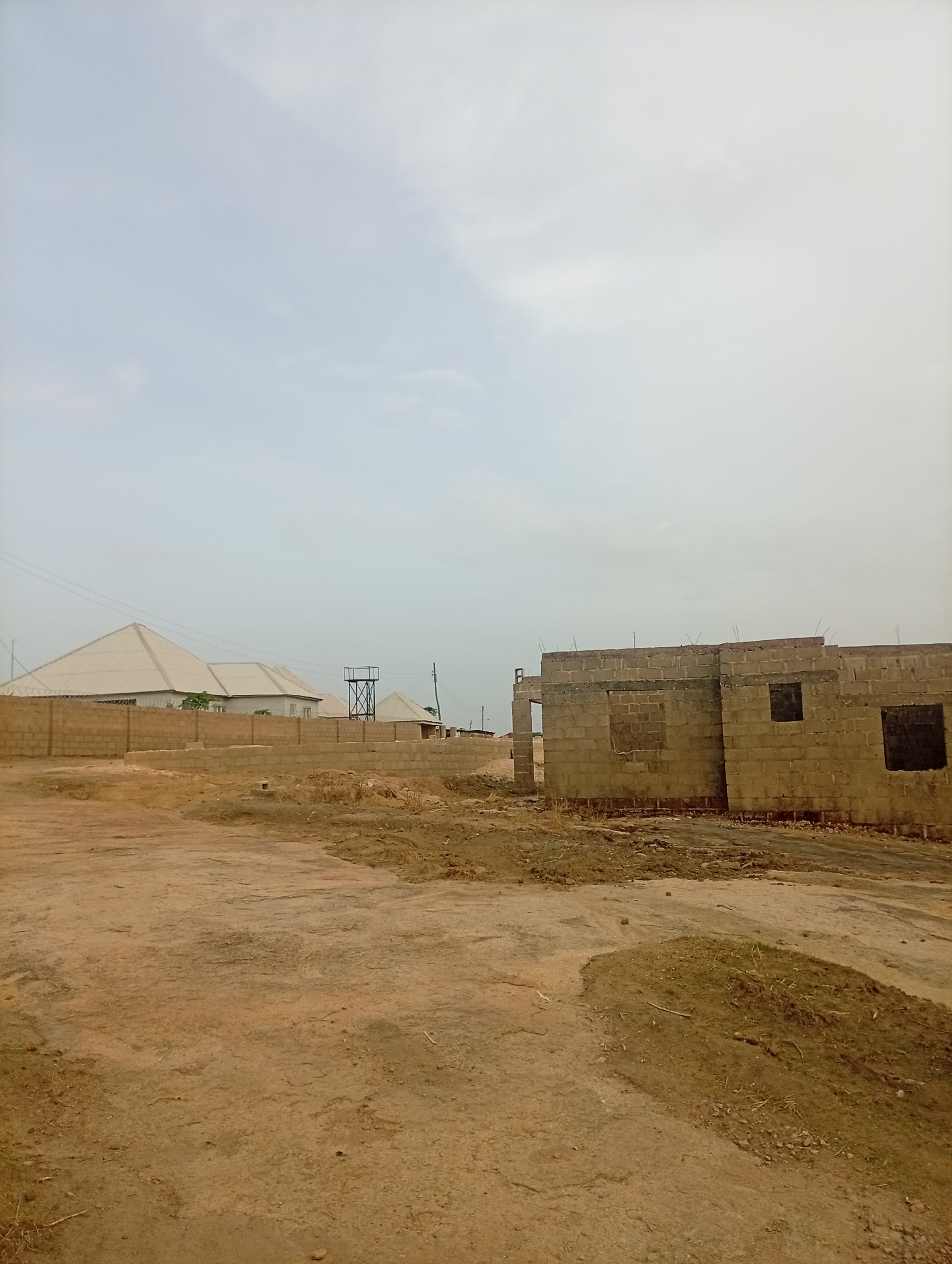 The image depicts a construction site in Jos, Nigeria. An unfinished building stands in the foreground, constructed from cinder blocks with window openings. In the background, completed structures are visible behind a brick wall. A water tower can be seen behind the completed buildings. The ground is predominantly bare earth, with patches of sparse vegetation. The sky is overcast. No people are visible in the frame.