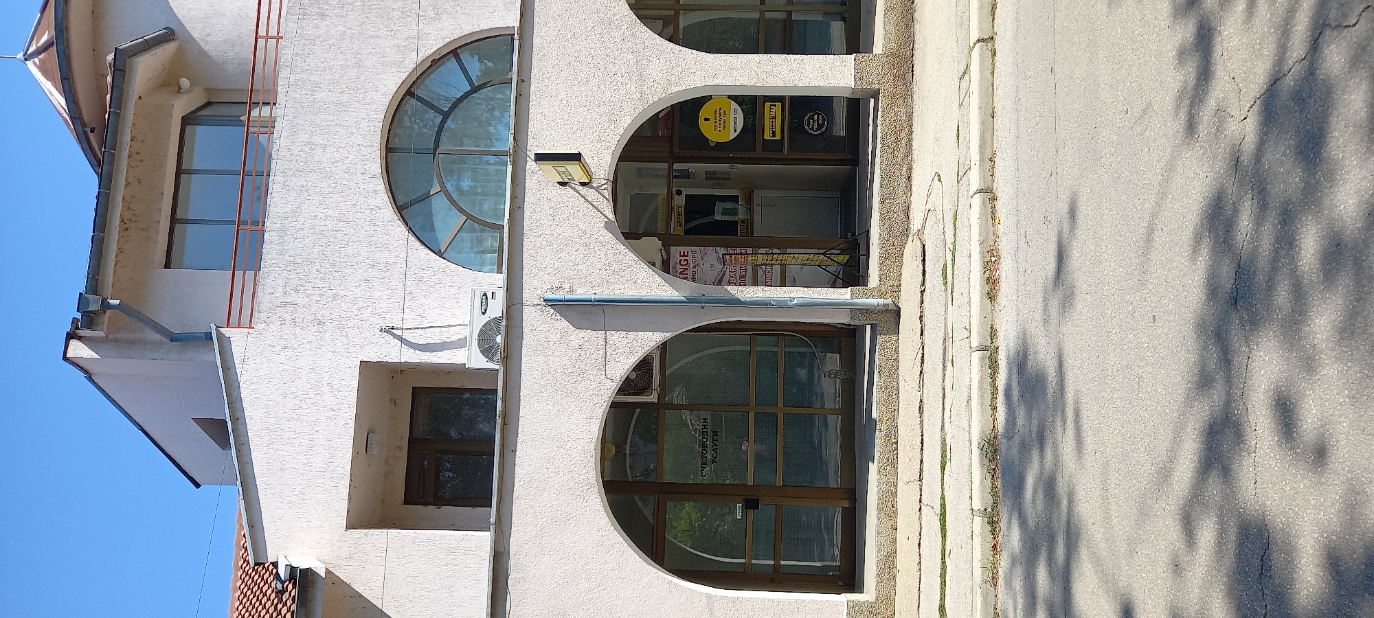 The image shows the facade of a building with arched entrances and windows, located in Silistra, Bulgaria. The building appears to be a commercial establishment. One of the ground-floor arches reveals a business with signage that includes the words "CHANGE" and "БИЗНЕС БЮРО", suggesting a currency exchange and business bureau. Another arch displays "СЧЕТОВОДНИ УСЛУГИ", indicating accounting services. There are several signs visible, including a yellow circular sign with an arrow and text, and a Western Union logo. An air conditioning unit is mounted on the wall above one of the ground-floor arches. The facade is textured and painted a light color, and the building has a roof with red tiles. A sidewalk and a street with shadows from trees are in the foreground. The sky is clear and blue. There are no people clearly visible in the image.