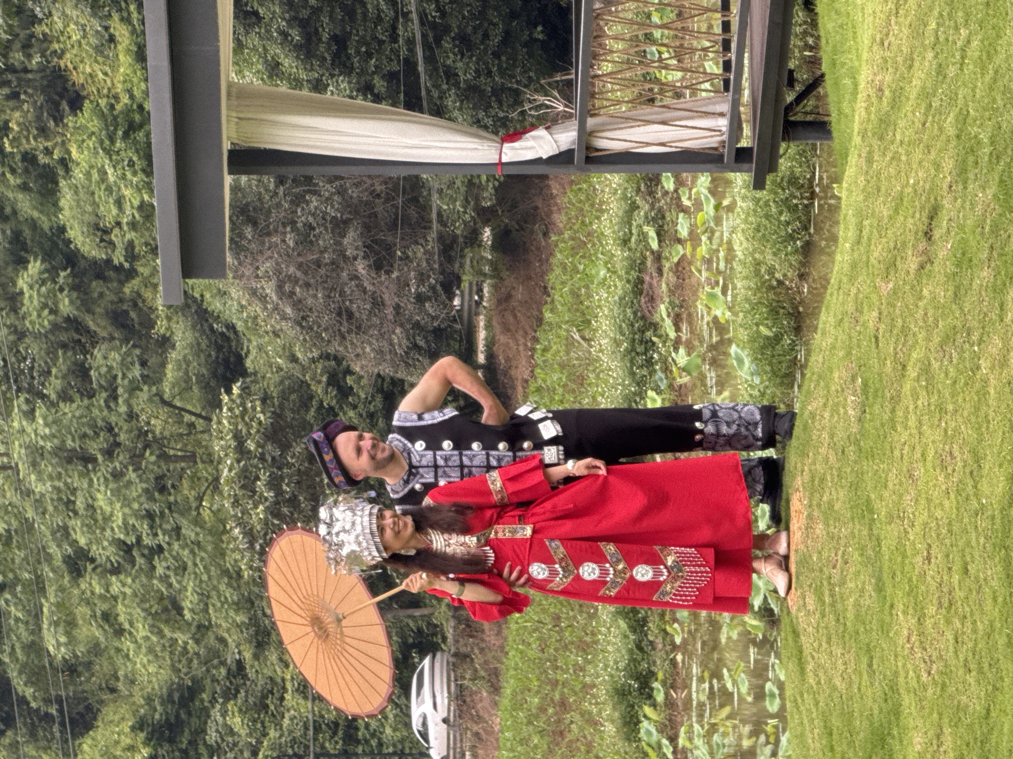 A medium shot shows two people in traditional clothing standing together in a grassy area of the Guilin, China area. A woman in a long red dress with elaborate gold embroidery is holding a yellow parasol, and she is looking off to the side, smiling. A man stands behind her, his hand on his hip. He wears a black and white patterned shirt and black pants. Both are wearing headwear. In the background is a pond with green plants, and a forest. Above them is a structure with a white curtain. A vehicle is slightly visible through the trees in the background.