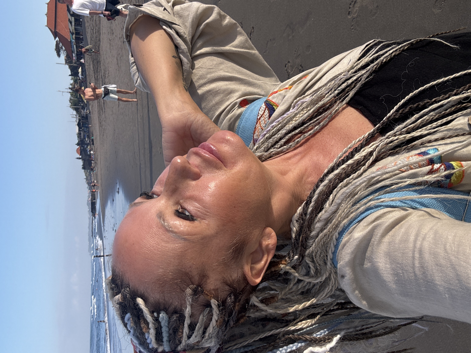 The image shows a close-up, selfie-style portrait of a woman with blonde and grey dreadlocks, taken on a beach in North Kuta, Indonesia. The woman is looking up and to her right, with her left arm bent and her hand resting on the back of her head. Her skin is shiny, likely from sweat or sunscreen, and she is wearing a light-colored, open jacket over a black top with a blue strap visible. The background shows a sandy beach with the ocean and waves on the left, and several people, beach umbrellas, and buildings, some with red roofs, further down the beach on the right. The sky is clear and blue. The overall impression is that of a warm, sunny day at a tropical beach.