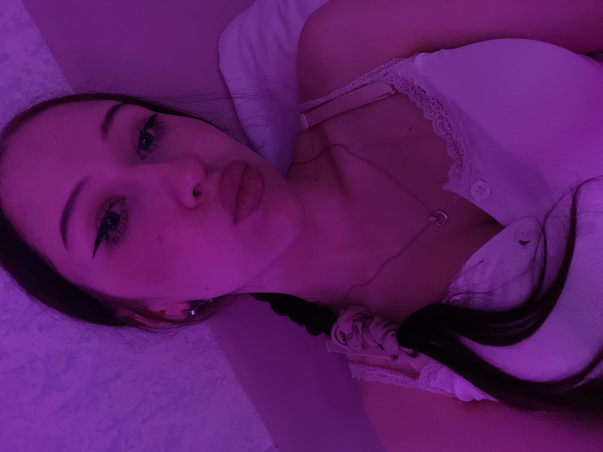 A young woman is lying in bed, illuminated by purple light. She is wearing a white camisole with lace trim and a heart-shaped necklace. Her hair is styled in a braid with a scrunchie. The background appears to be a wall with a textured surface. The image is a close-up selfie taken from a high angle. There is no information about the location in Votkinsk, Russia.