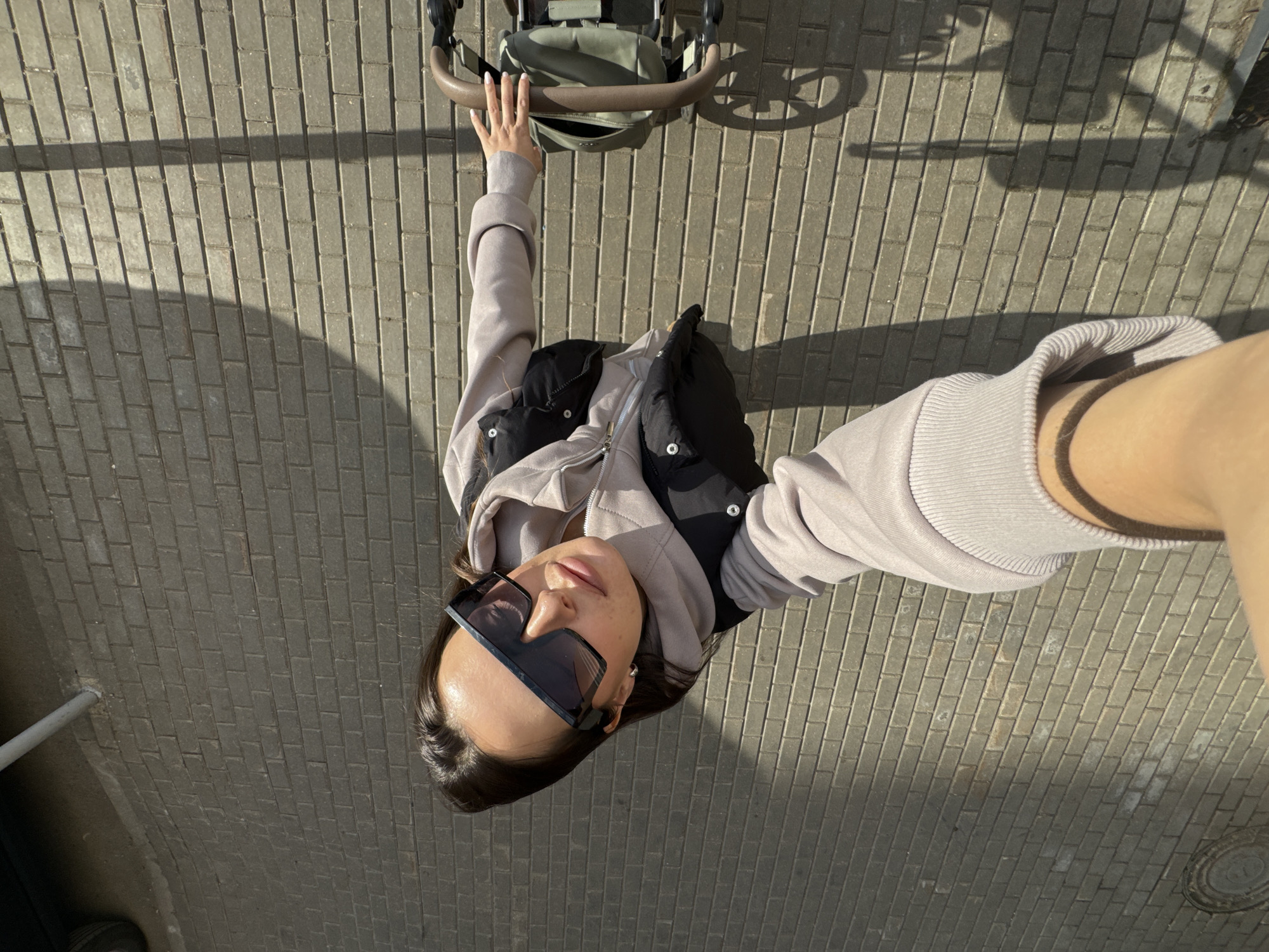 An overhead-view photo shows a person standing outdoors on a sunny day in what is contextually Akulovo (Gp Kubinka), Russia. The subject is a female with dark hair, wearing large, dark rectangular sunglasses that obscure her eyes. She is dressed in a light-colored (possibly beige or light grey) hooded sweatshirt with a black puffy vest layered over it.
Her left hand rests on the brown handle of an olive-green stroller or pram, which is positioned in front of her. The photo is taken from a high angle, likely a selfie, with her right arm and hand holding the camera visible on the far right of the frame. Her face is turned upwards towards the camera, illuminated by bright sunlight.
The ground beneath her is paved with grey rectangular bricks, arranged in a diagonal pattern. Strong, elongated shadows stretch across the paving from left to right, indicating bright sunshine and a clear sky. Notable shadows include a parallel set of lines resembling a fence or railing on the left and a circular shape, possibly from a wheel, on the right. No visible text is present in the image. The overall impression is a casual, everyday scene captured in good weather.