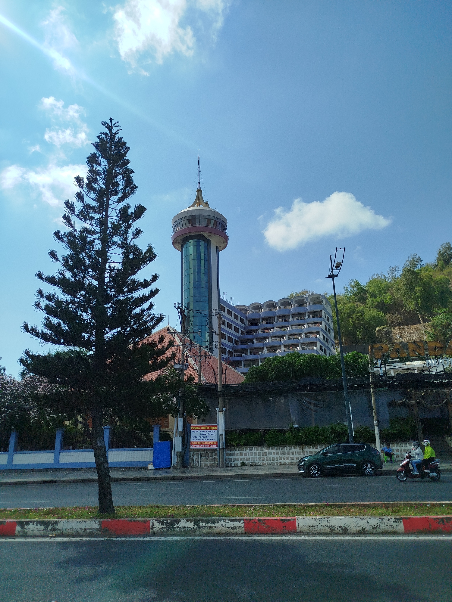 The image presents a cityscape with a notable observation tower in the background. A tall, dark green pine tree is prominently positioned on the left side of the frame, contrasting with the light blue sky and scattered clouds.
Behind the tree, the observation tower stands out with its cylindrical shape and golden dome-like top. To the right of the tower, a multi-story building with balconies is visible, and further to the right, a hillside covered in green vegetation completes the backdrop.
A road runs across the bottom of the image, marked by a curb painted with red and white stripes. On the sidewalk adjacent to the road, a dark green SUV is parked. A motorcycle with two people wearing helmets and jackets is moving along the road.
A utility pole is located near the parked car. A recruitment sign with Vietnamese text can be seen on a building close to the tree on the left.
Based on the architectural style of the tower and the vegetation, this could potentially be Dalat City, Vietnam.