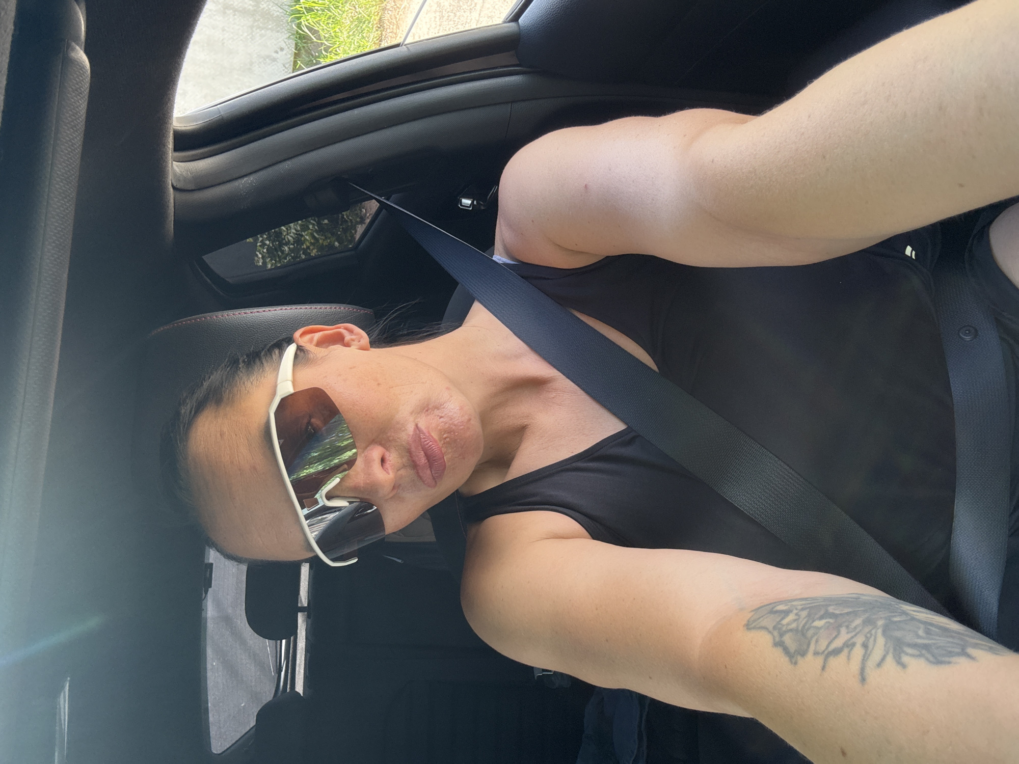 In a car, a woman with sunglasses and a black tank top is taking a selfie. She has a tattoo on her left arm. The seatbelt is visible across her chest. The car's interior is dark, with a bright reflection on the sunglasses, indicating an outdoor setting. The background shows greenery and a light-colored wall, suggesting a residential or natural area. The image appears to be taken during the daytime. The exact location in Chaikovsky, Russia, cannot be determined from the visual information alone.