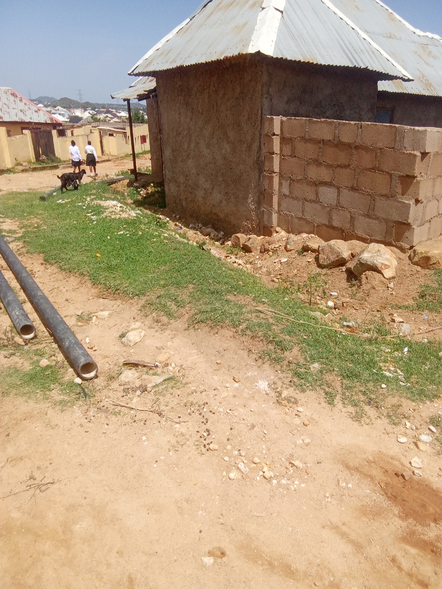 In Barakin Akawo, Nigeria, two individuals wearing white tops and dark shorts walk on a dirt path near a building constructed of earth and a brick wall. A dark-colored dog follows them. Several black pipes are laid out on the ground in the foreground. The environment appears arid, with sparse vegetation and some scattered rocks. The sky is clear and blue.