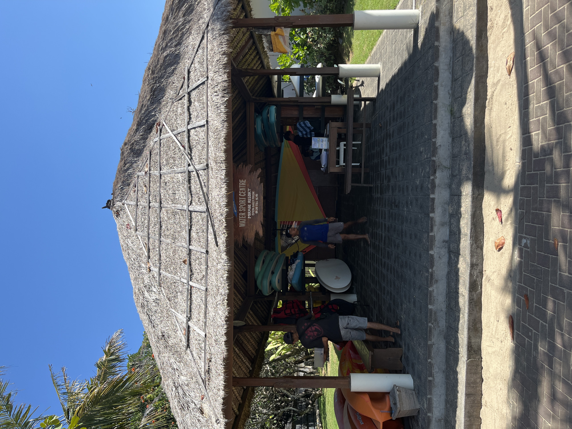 The image depicts a water sport center in Kuta Selatan, Indonesia, characterized by its thatched-roof pavilion. The pavilion houses various water sports equipment, including kayaks, surfboards, life vests, and paddleboards, neatly organized on shelves and leaning against the structure. Two men are present; one is standing inside the pavilion, appearing to interact with a desk, while the other is walking away from the pavilion carrying a white rectangular object. The ground is paved with dark tiles, and the surrounding area features lush green grass and palm trees, indicating a tropical environment. The sky is clear blue, suggesting a sunny day.