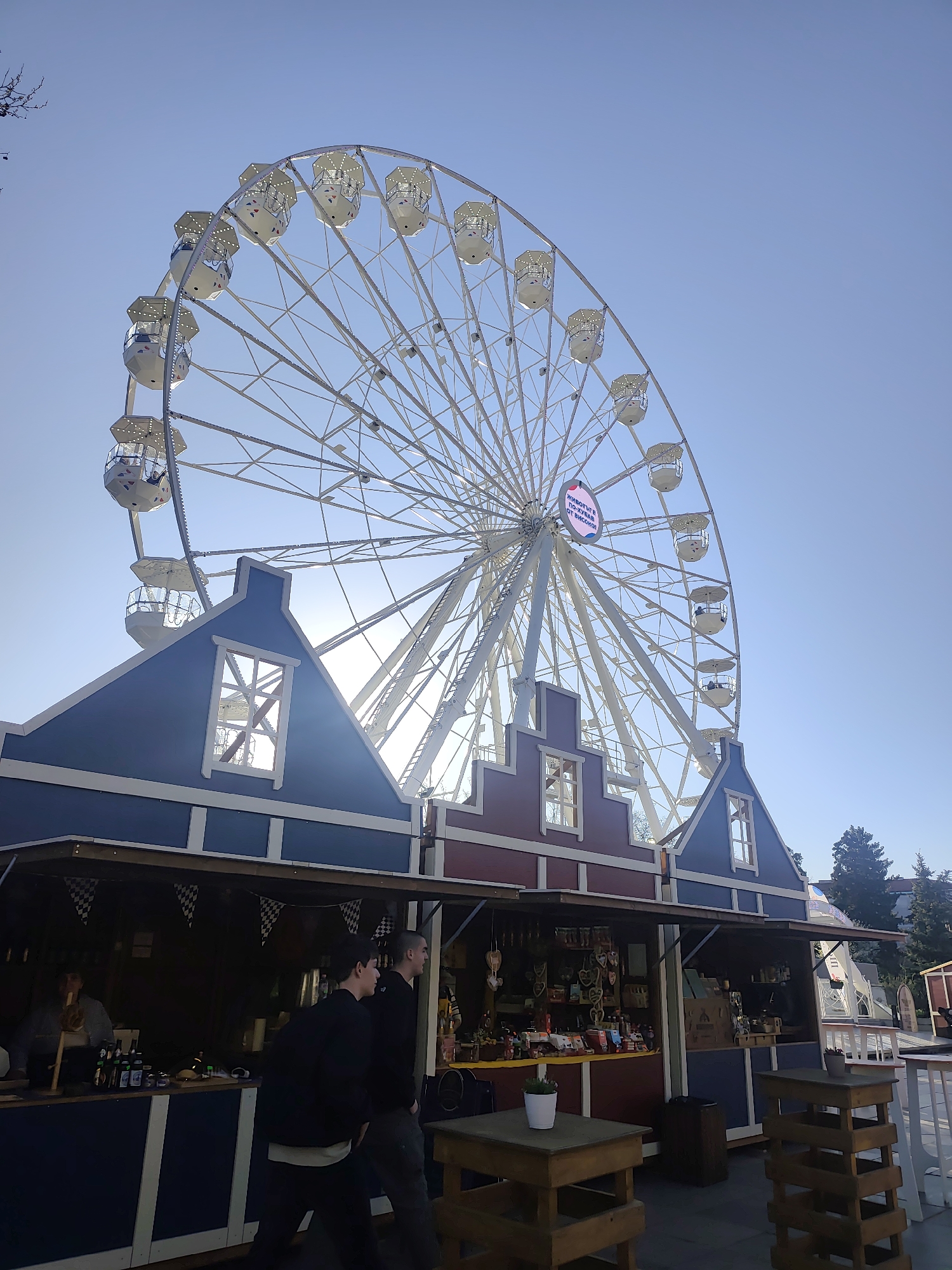 The image depicts a Ferris wheel standing behind market stalls on a clear day. The Ferris wheel is white, with individual cabins. One side has a pink circular sign on it. The market stalls are painted in blue and red, styled to resemble traditional European houses. Two men wearing dark clothing are walking in front of the stalls. There are wooden tables and stools in front of the stalls. This is located in the city of Sofia, Bulgaria.