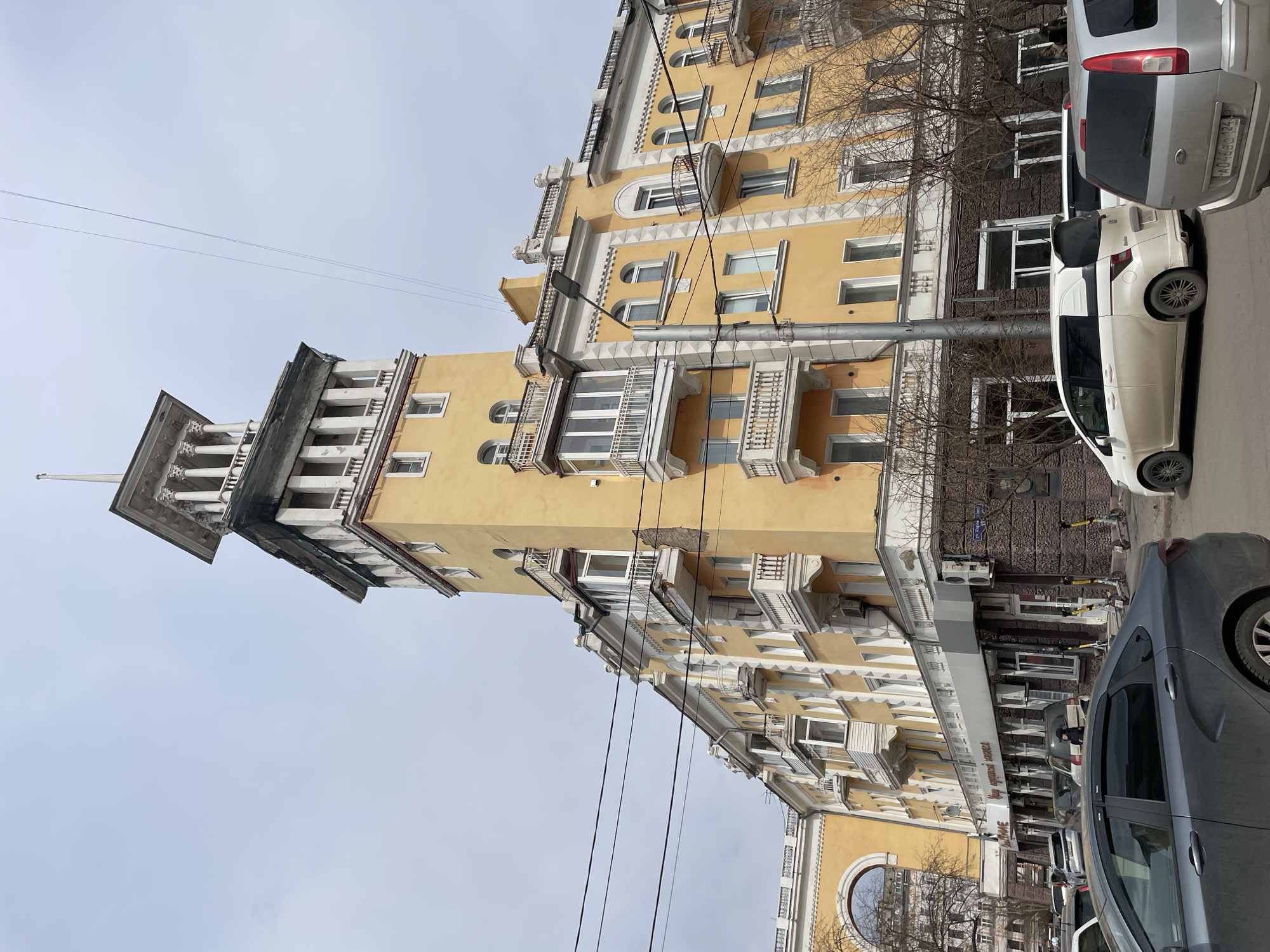 The image captures a section of Mira Avenue in Krasnoyarsk, Russia. The focus is on a yellow architectural building, likely an apartment complex or commercial building, with white trim and detailing. The building includes a prominent tower feature with a pointed roof. Power lines run across the facade of the building, crossing the sky. Parked cars are visible in the foreground, partially obstructing the view of the building's base. The sky is overcast, suggesting a cloudy day. There are some trees with no leaves are visible in front of the building. A street sign is mounted along the side of the building. A silver minivan and white hatchback car are present.