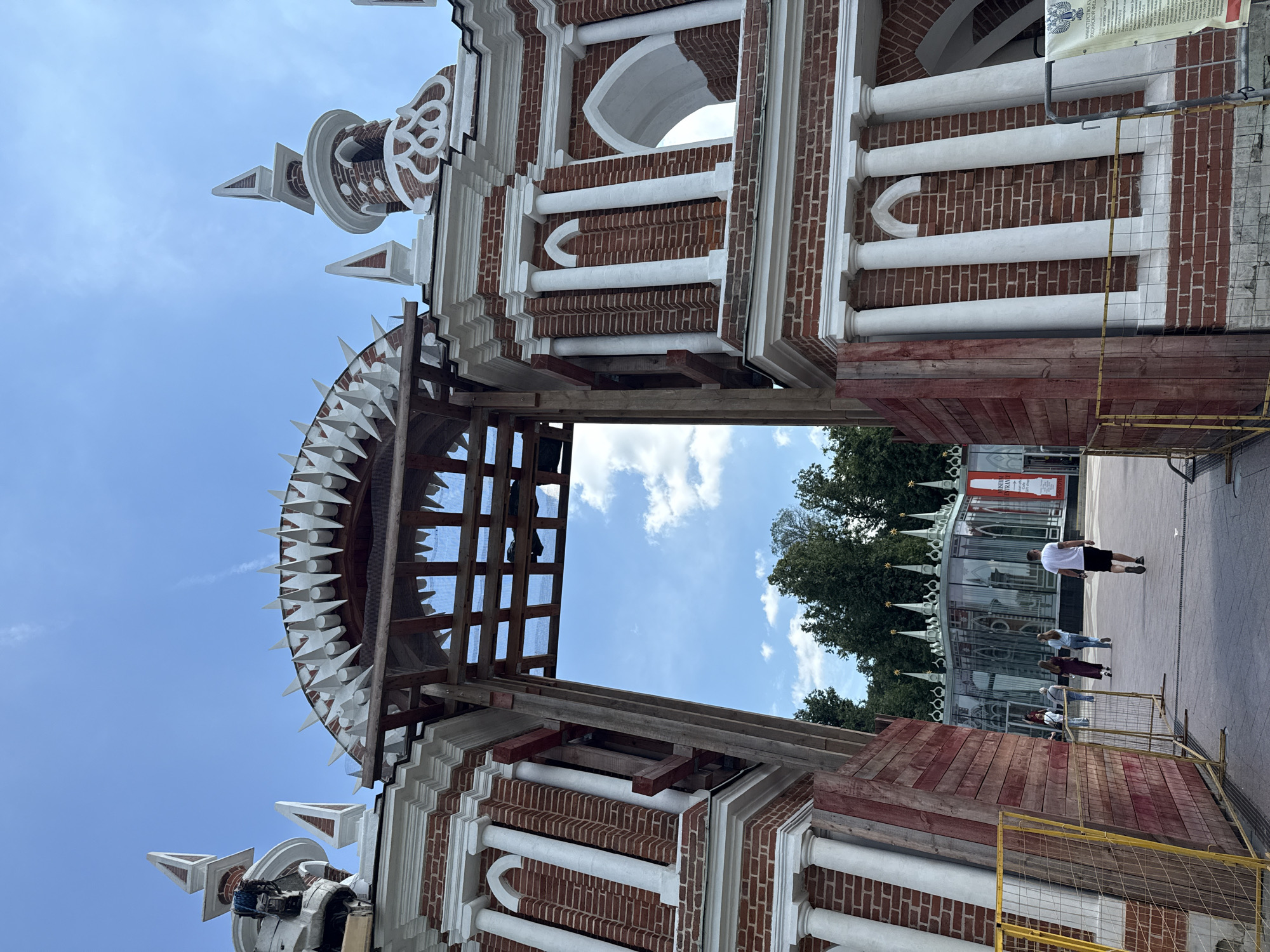 The image depicts an outdoor scene in Moscow, Russia, showcasing a section of an ornate brick building with white architectural details. The building features classical columns, decorative brickwork, and pointed spires. Parts of the structure appear to be under renovation or construction, with visible wooden scaffolding and protective barriers.
In the background, a modern glass structure with similar pointed spires can be seen, possibly part of the same complex. Trees line the edge of the scene, suggesting a park or garden setting. The sky is a clear blue with scattered white clouds, indicating fair weather.
A few people are present in the scene, walking along a paved pathway. One person in a white t-shirt and black shorts is walking away from the camera, while others are further in the distance. The overall atmosphere seems casual and public, consistent with a tourist area or a historical site.
A yellow construction fence and a wooden barrier are prominent in the foreground, further emphasizing the ongoing work on the building. A sign with Russian text is partially visible on a pole to the right of the image. The time of day appears to be daytime, with bright lighting.