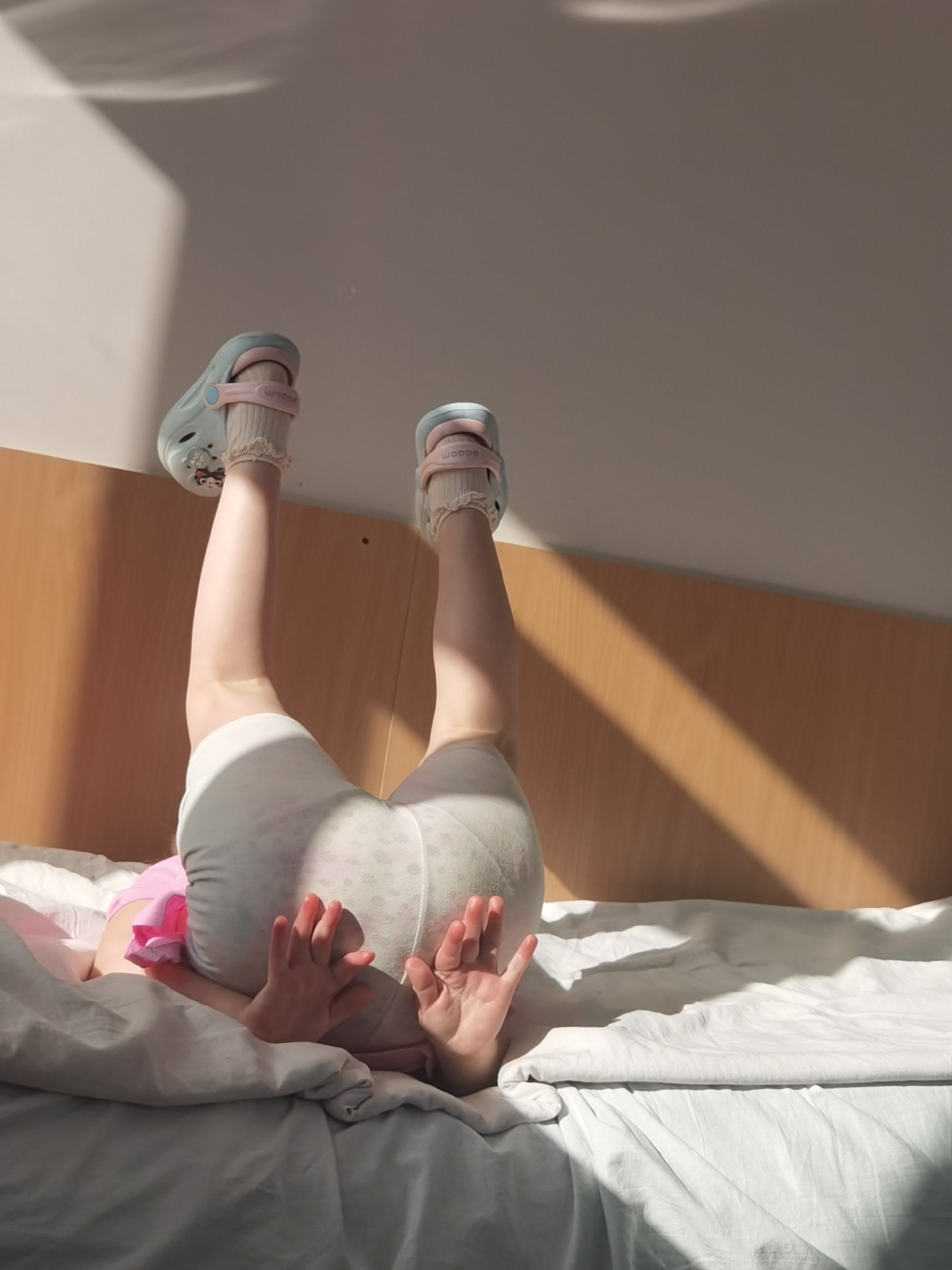A child is lying on a bed in what appears to be a room, with their legs straight up in the air. The child is wearing light-blue Crocs with pink straps and white socks with lace trim. They are also wearing white shorts. The child's hands are visible, reaching out from behind a gray pillow. The bed has white sheets, and there is a light-pink shirt visible. A wooden headboard is behind the bed, and the wall is a light color. Light is streaming in, creating shadows on the wall. The image was taken in Perm, Russia.