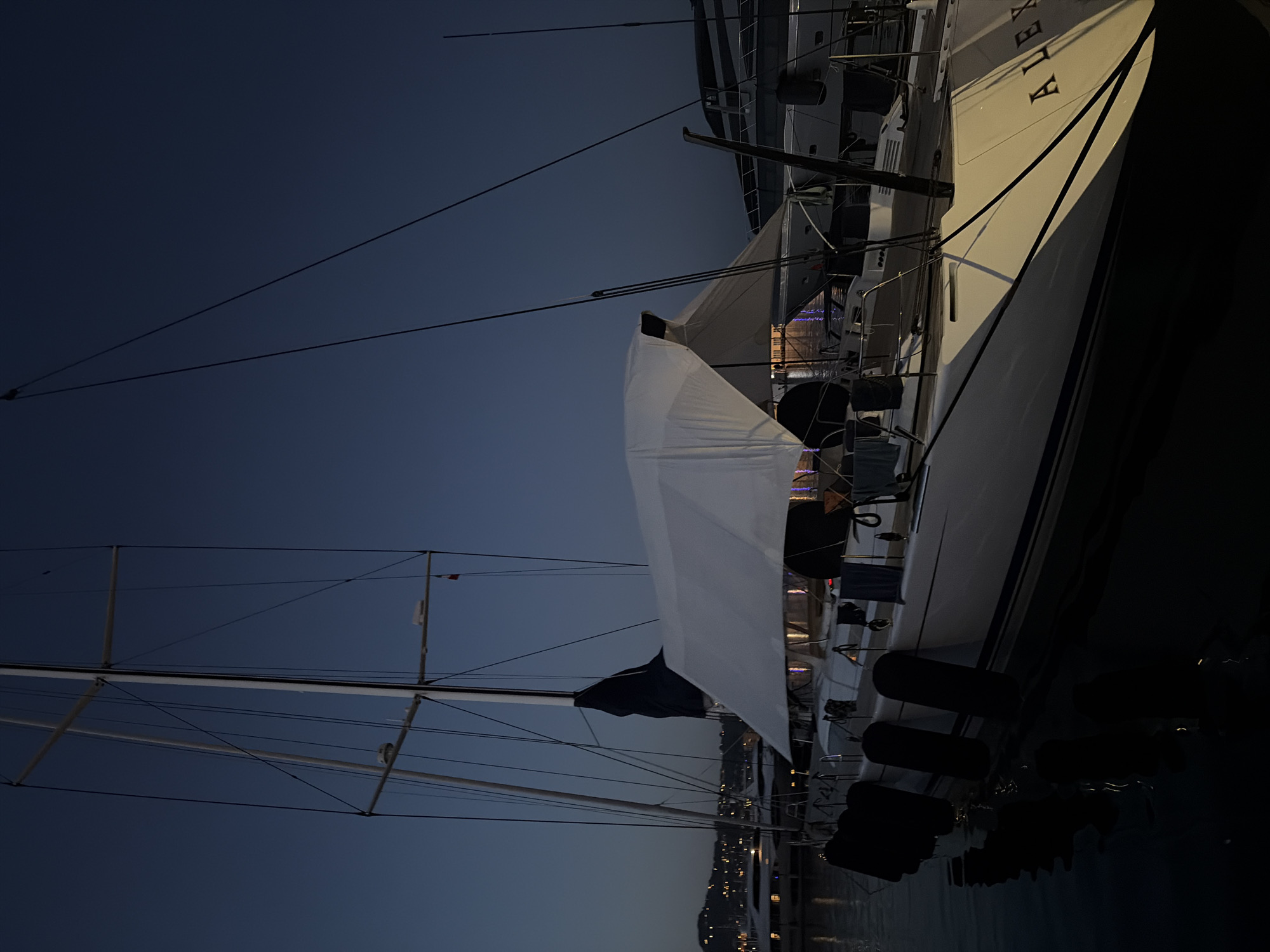 A sailboat named "ALEX" is docked at night in the city of Imperia, Italy. The sailboat has a white hull and a dark blue sail furled on the mast. A white tarp is stretched over the boom, creating a makeshift shelter on the deck. The boat is equipped with several black fenders hanging along its side. In the background, other boats and distant city lights are visible. The water appears dark and still, reflecting the faint lights. The sky is a deep blue, indicating twilight or early night. There are no people visible in the image.