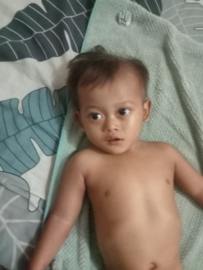 A shirtless toddler lies on a light green towel, placed atop a bed with a blue and white leafy pattern. The child has dark hair, big eyes, and slightly tanned skin. The focus is on the child's face and upper body. The background includes the patterned bedding and the folded towel providing a soft, textured surface beneath the child.