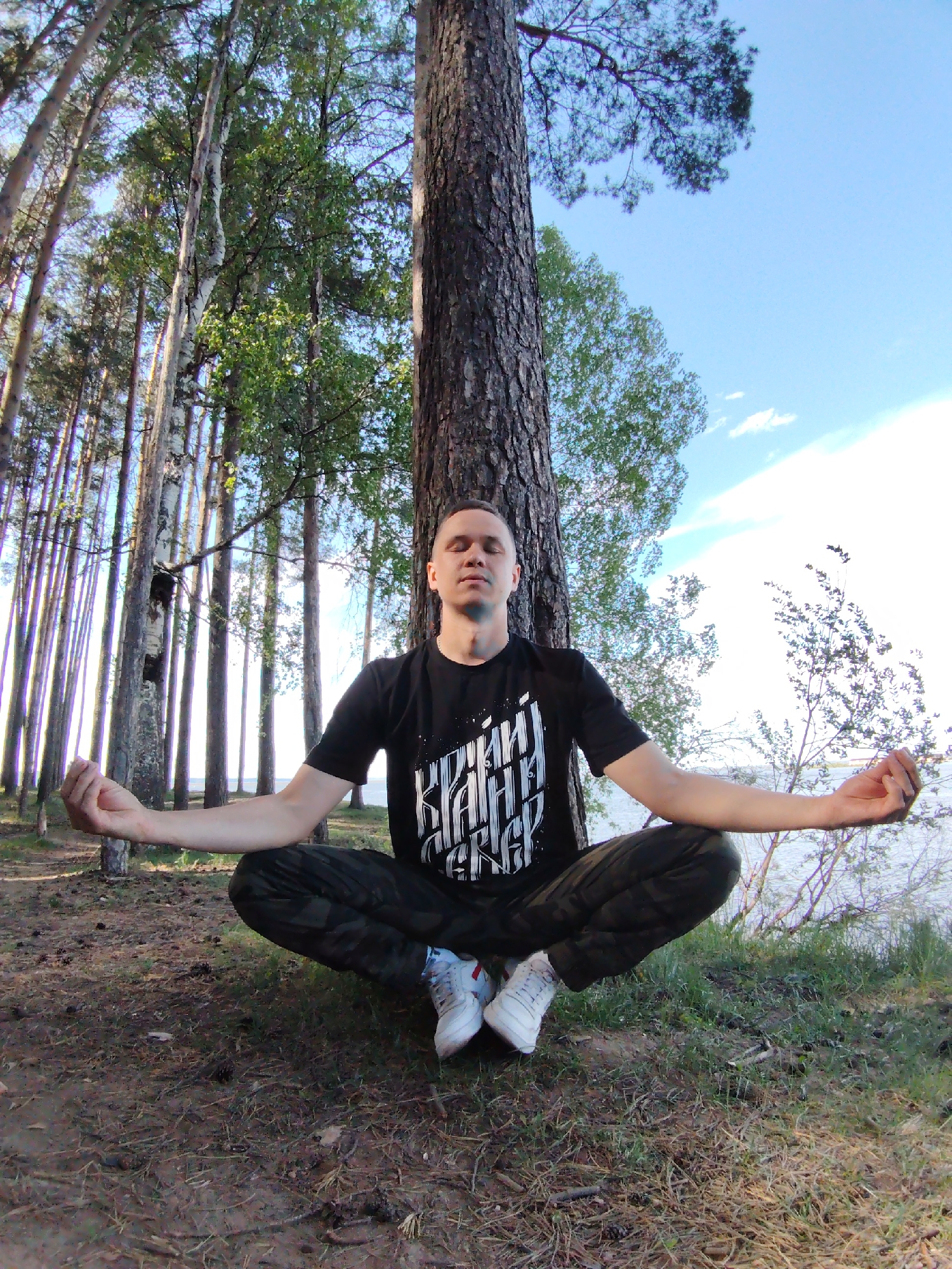 A man sits in a meditative pose in front of a tree in a forest. The location is Chaikovsky, Russia. The man is wearing a black t-shirt with white Cyrillic text, camouflage pants, and white sneakers. He is sitting cross-legged with his hands outstretched in a meditative gesture. His eyes are closed. A tall, thick-trunked tree is directly behind him. The forest consists of a mix of birch and pine trees. The ground is covered in pine needles and grass. In the background, a body of water is visible, with trees and bushes along the shoreline. The sky is blue with some clouds.